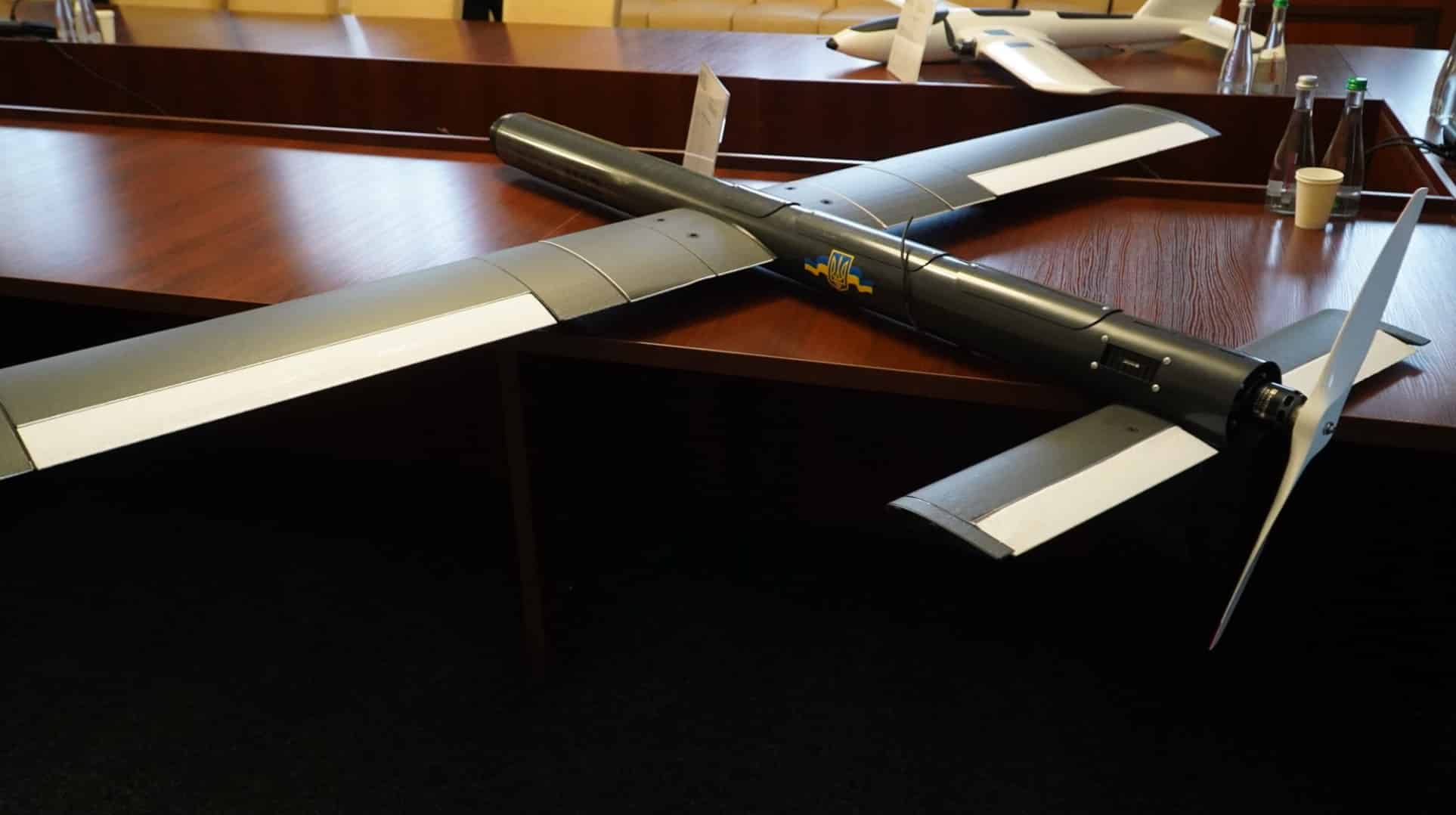 Olive fixed-wing kamikaze drone with swept wings resting on a conference room table.