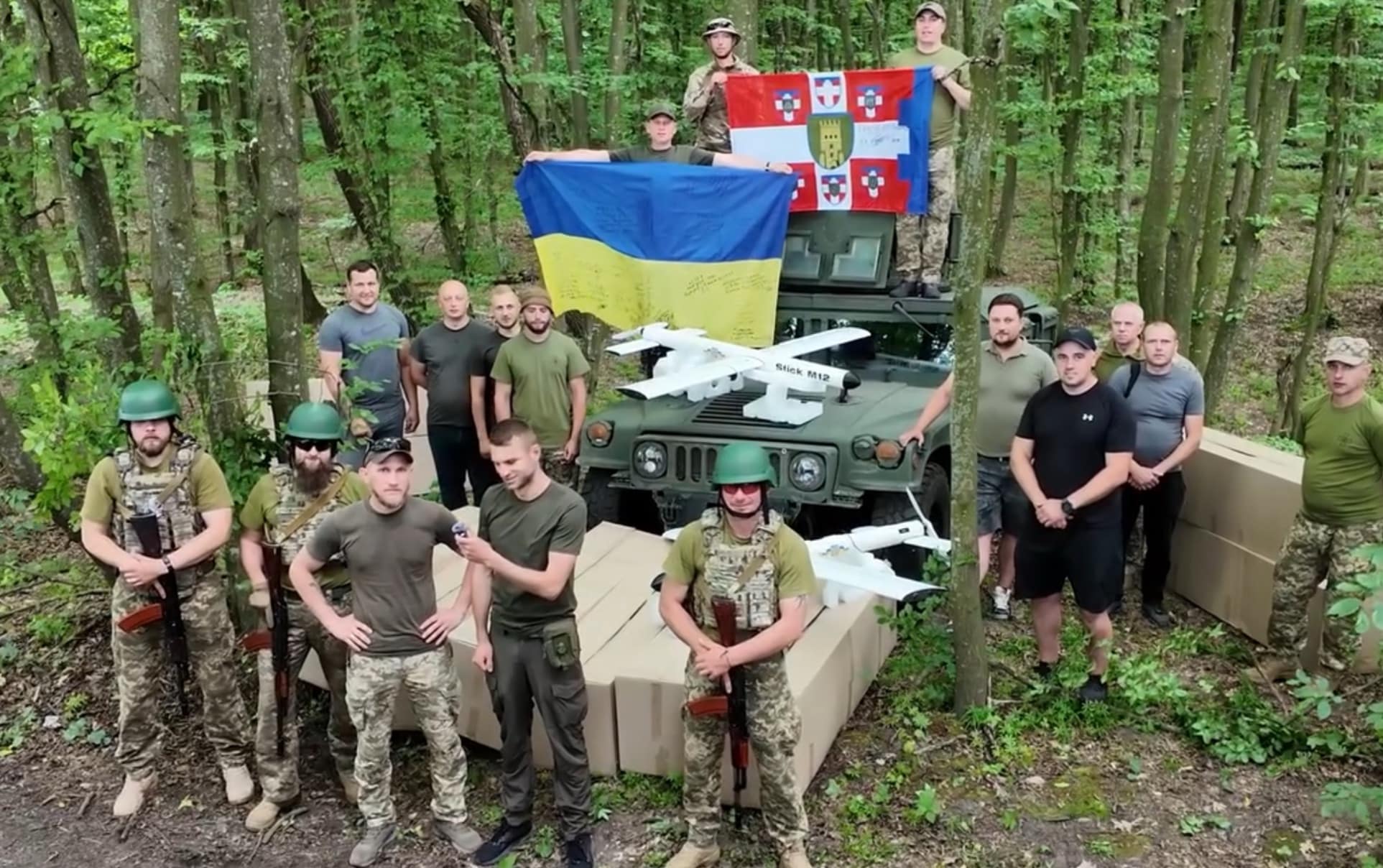 Ukrainian soldiers pose in woodland with a white fixed-wing drone and unit flags.