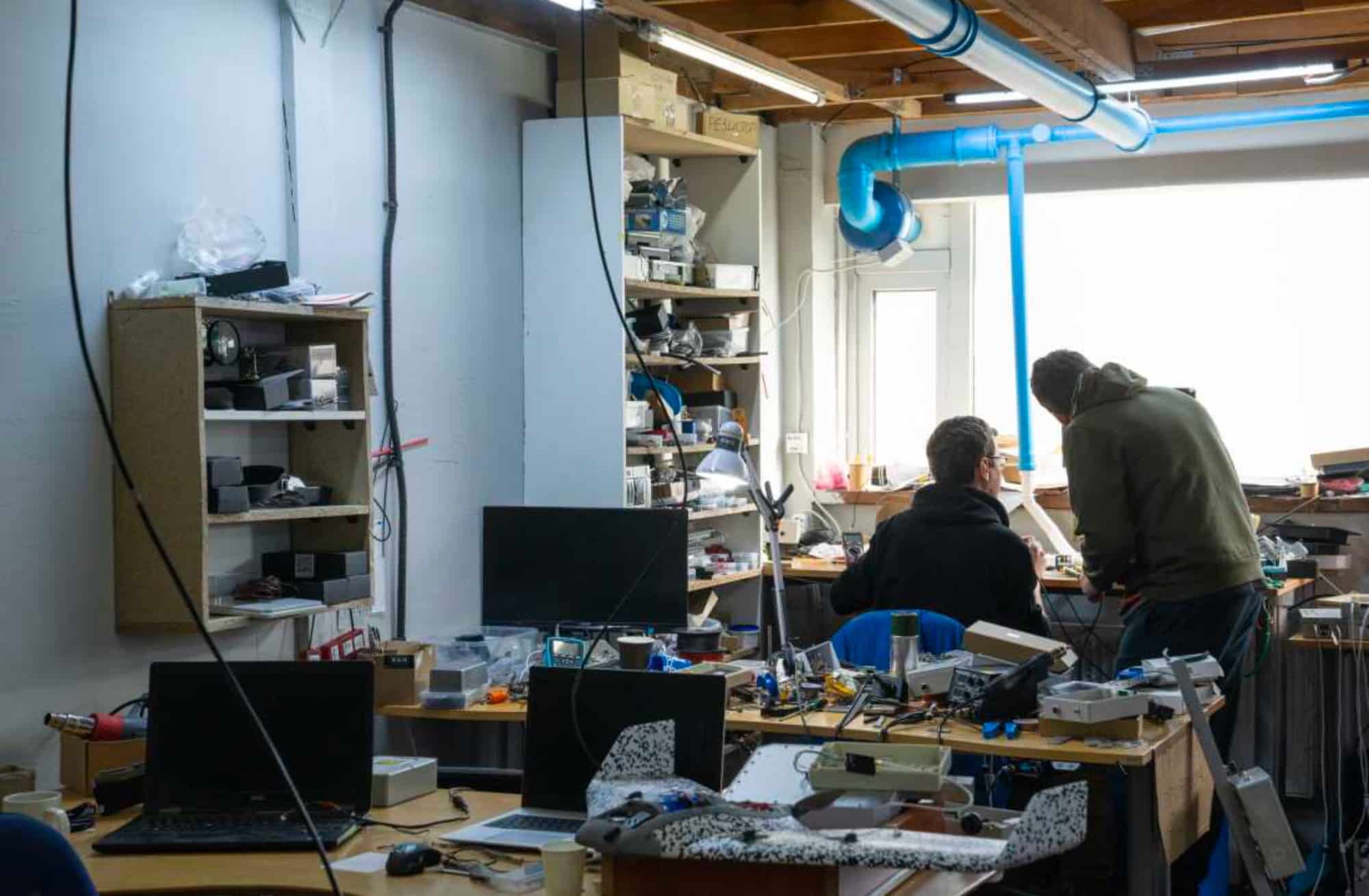 Two workers assemble drone electronics at cluttered benches in a small workshop.