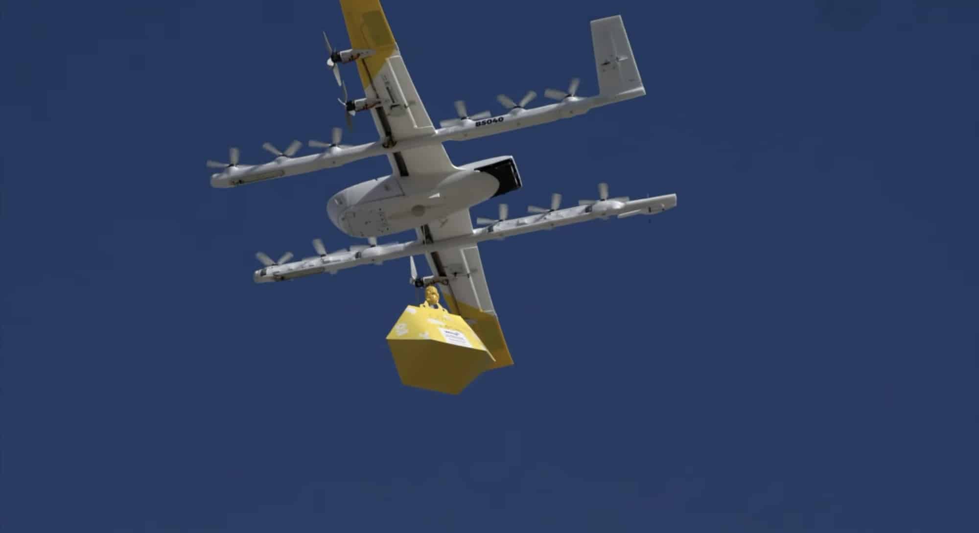 A Wing delivery aircraft hovers with a yellow package clipped beneath its fuselage.