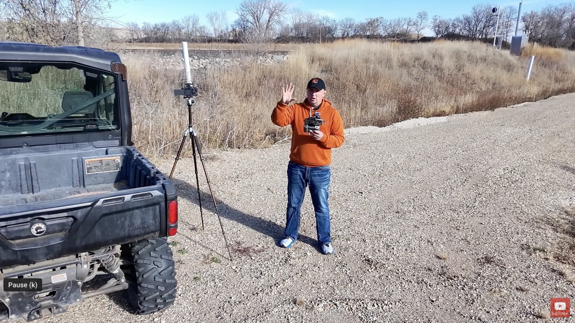 Pilot in an orange hoodie holds a DJI controller beside a UTV on gravel.