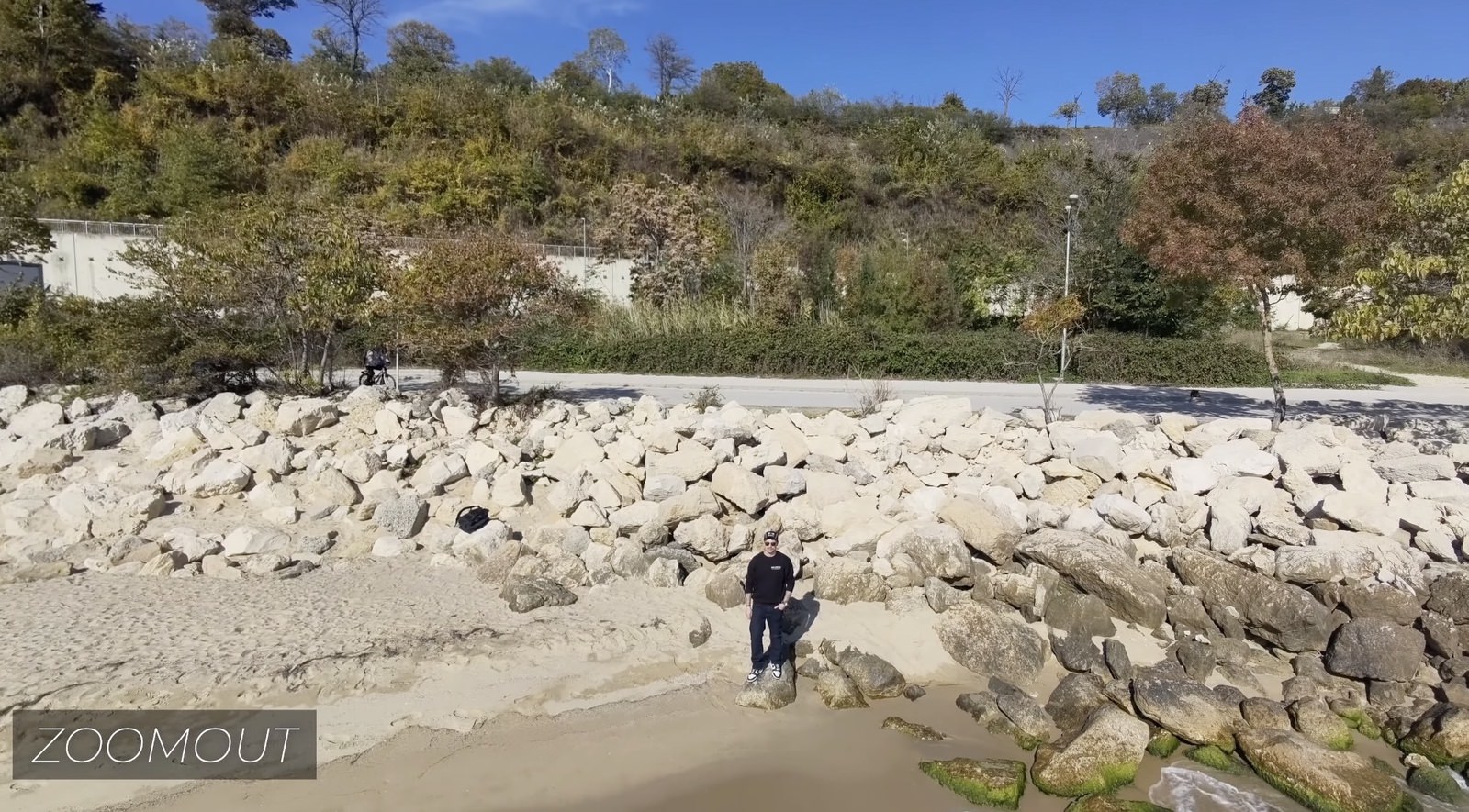 Aerial zoom-out frame of a lone figure on a boulder-strewn shoreline below wooded bluffs.
