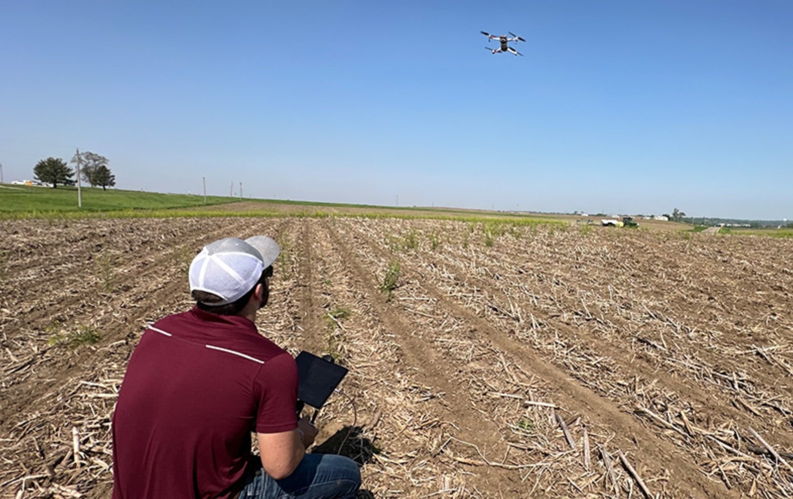 Student kneels at the edge of a harvested field flying a small drone.