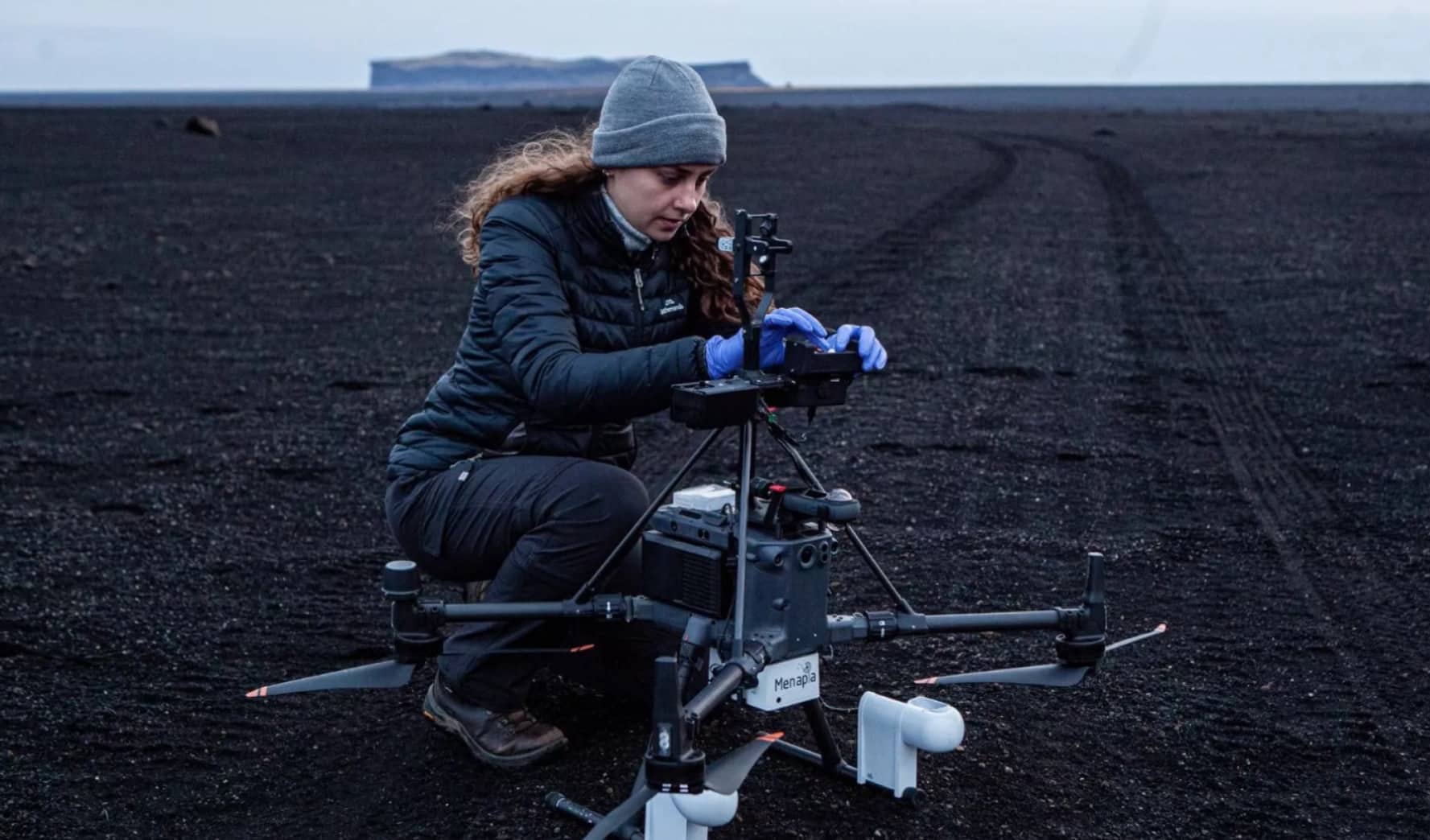 Researcher kneeling on black volcanic sand preparing a sensor-equipped drone in Iceland.