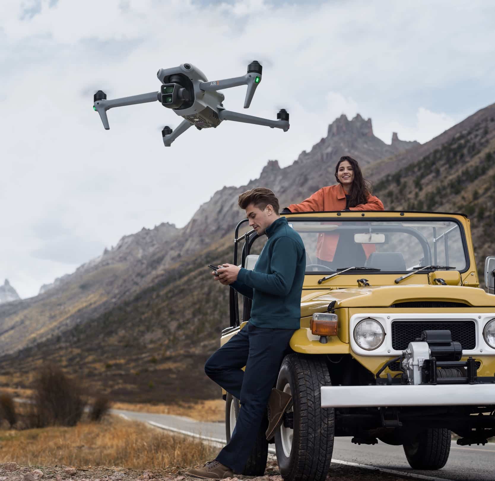 DJI Air 3 hovering beside two people and a yellow off-road vehicle in mountains.