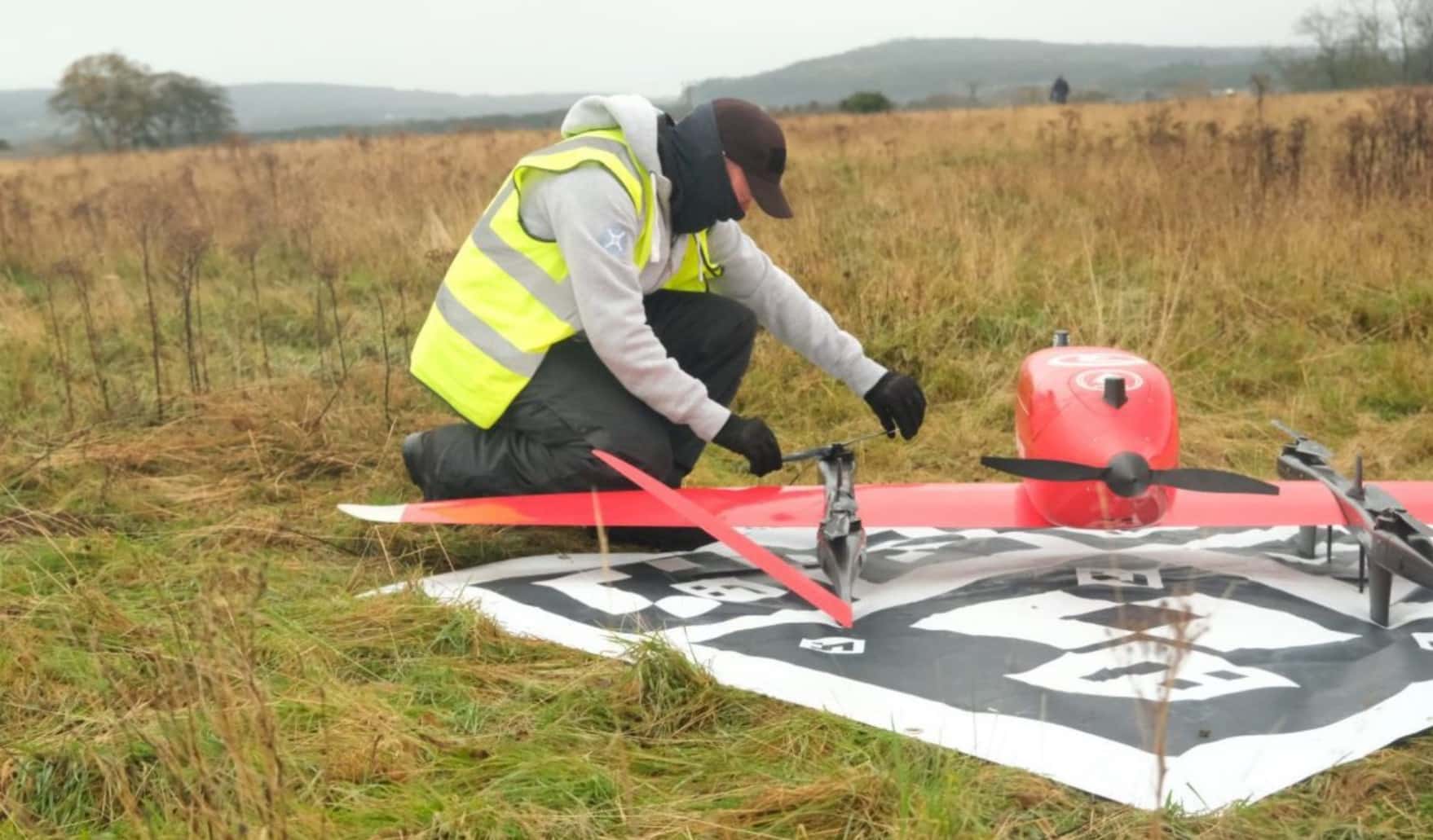 Operator in hi-vis kneeling beside a red and white delivery drone in a field.