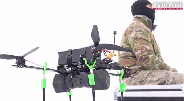 Soldier in a balaclava sits beside a large payload-carrying multirotor drone.