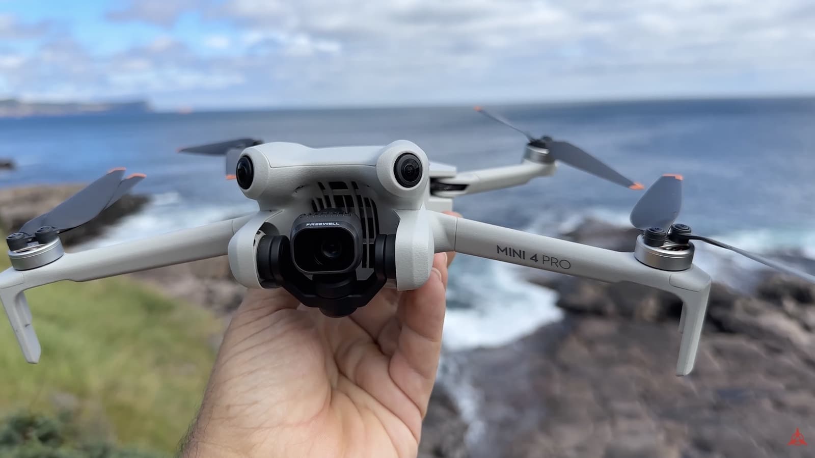A hand holds a DJI Mini 4 Pro above a rocky coastline.