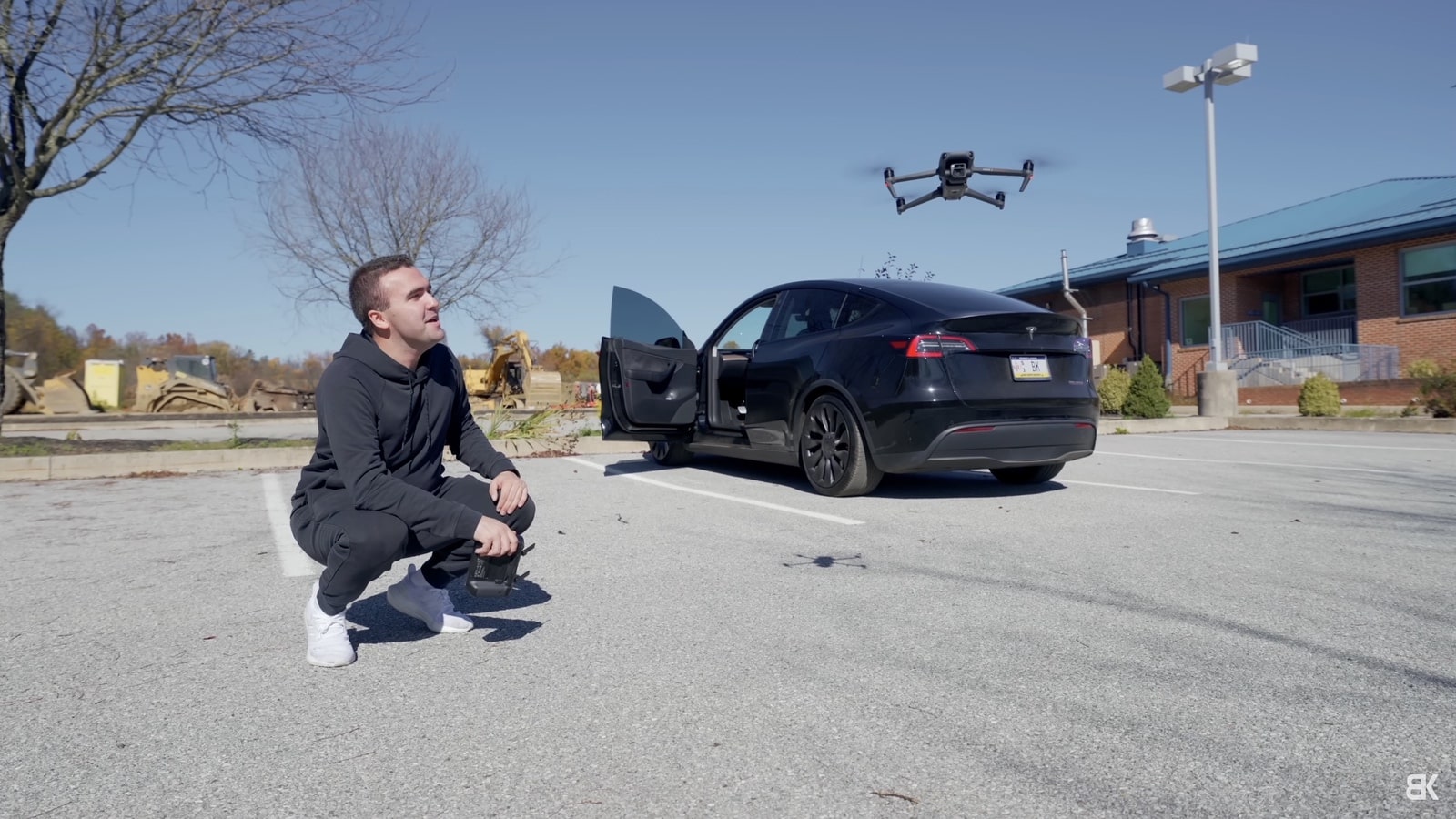 A pilot crouches in a lot as a drone hovers above his open car.