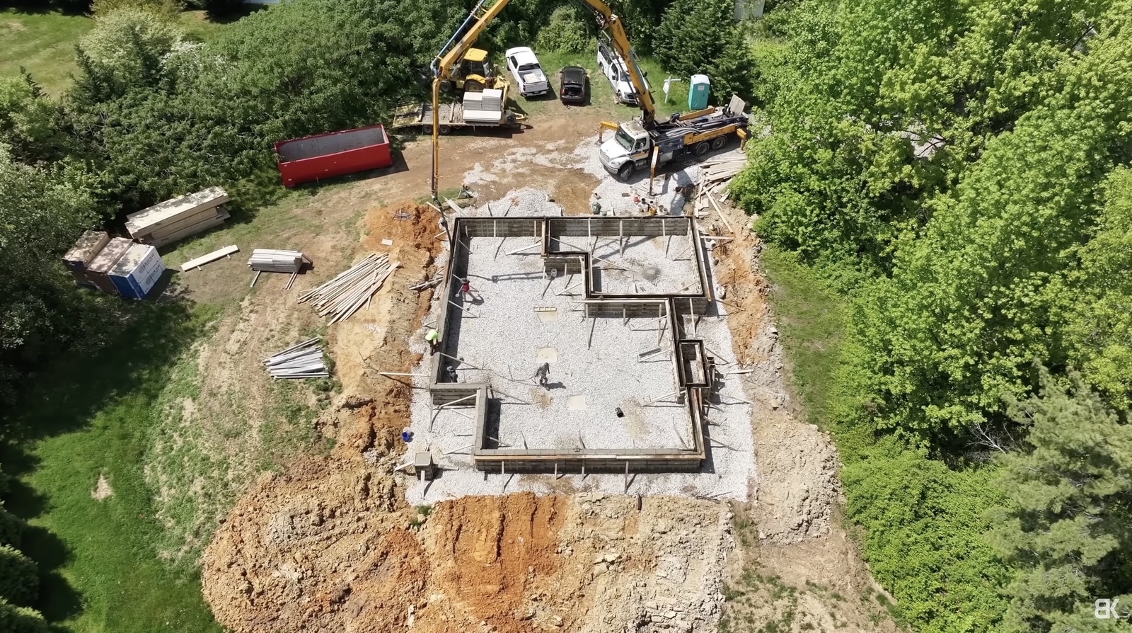 Aerial view of a poured foundation and excavators on a wooded building site.