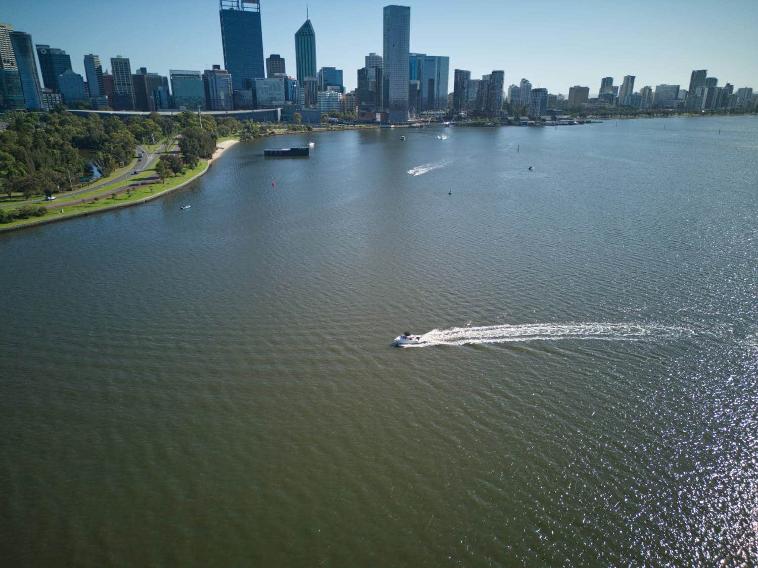 Aerial over a wide river toward a city skyline with a speedboat trailing a wake.