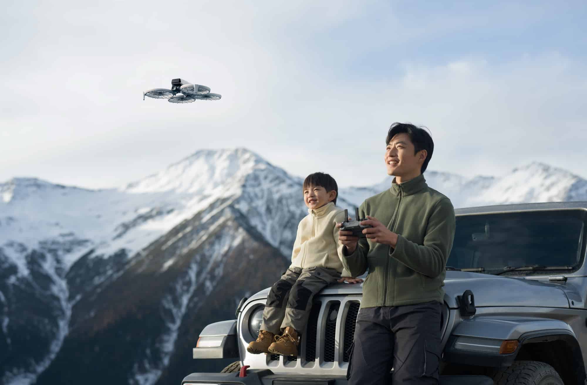 Man and boy on a Jeep hood watching the Flip against snowy peaks.
