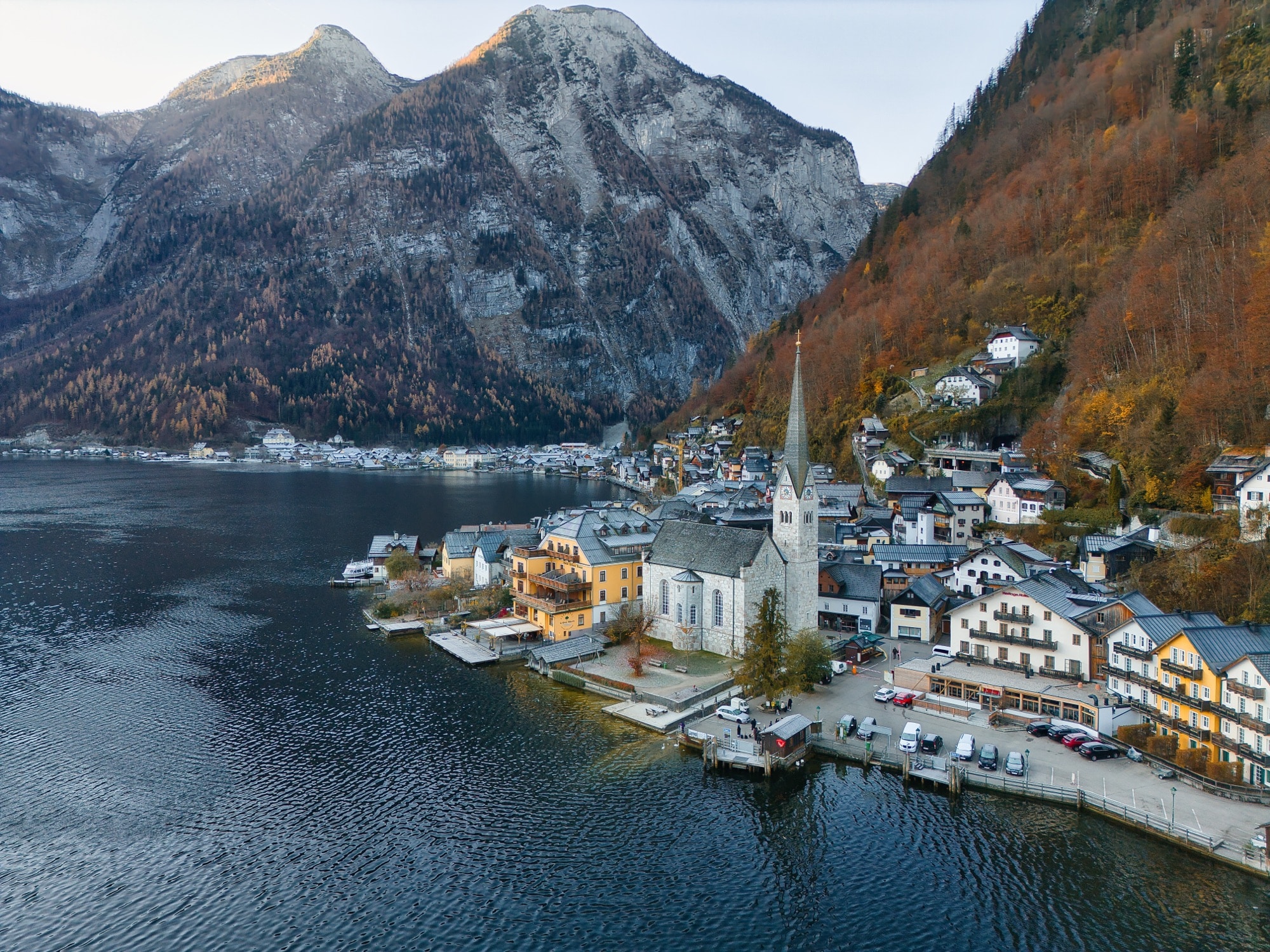 Aerial view of Hallstatt's lakeside church and houses below steep autumn mountains.