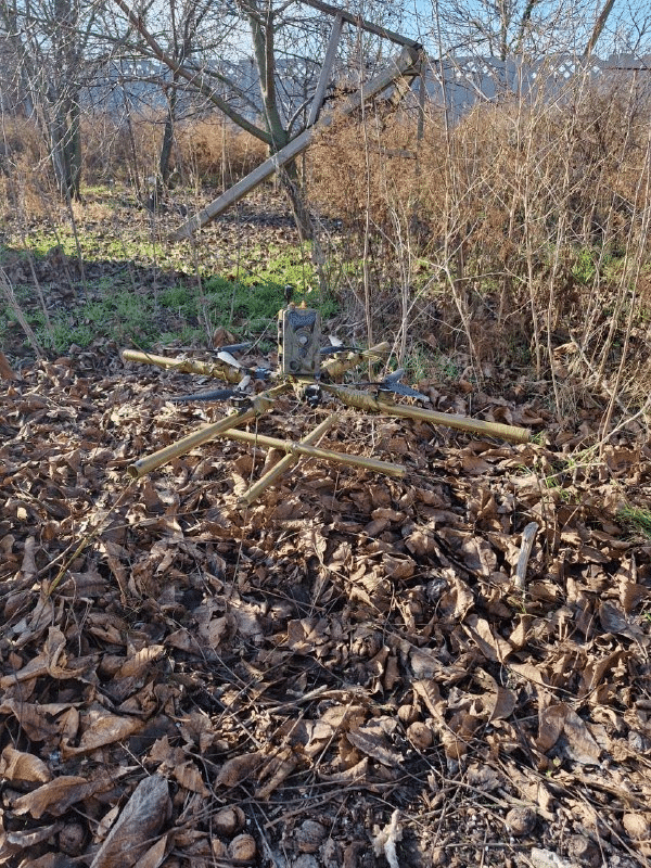 Camouflaged FPV drone fitted with a trail camera resting among dead leaves.