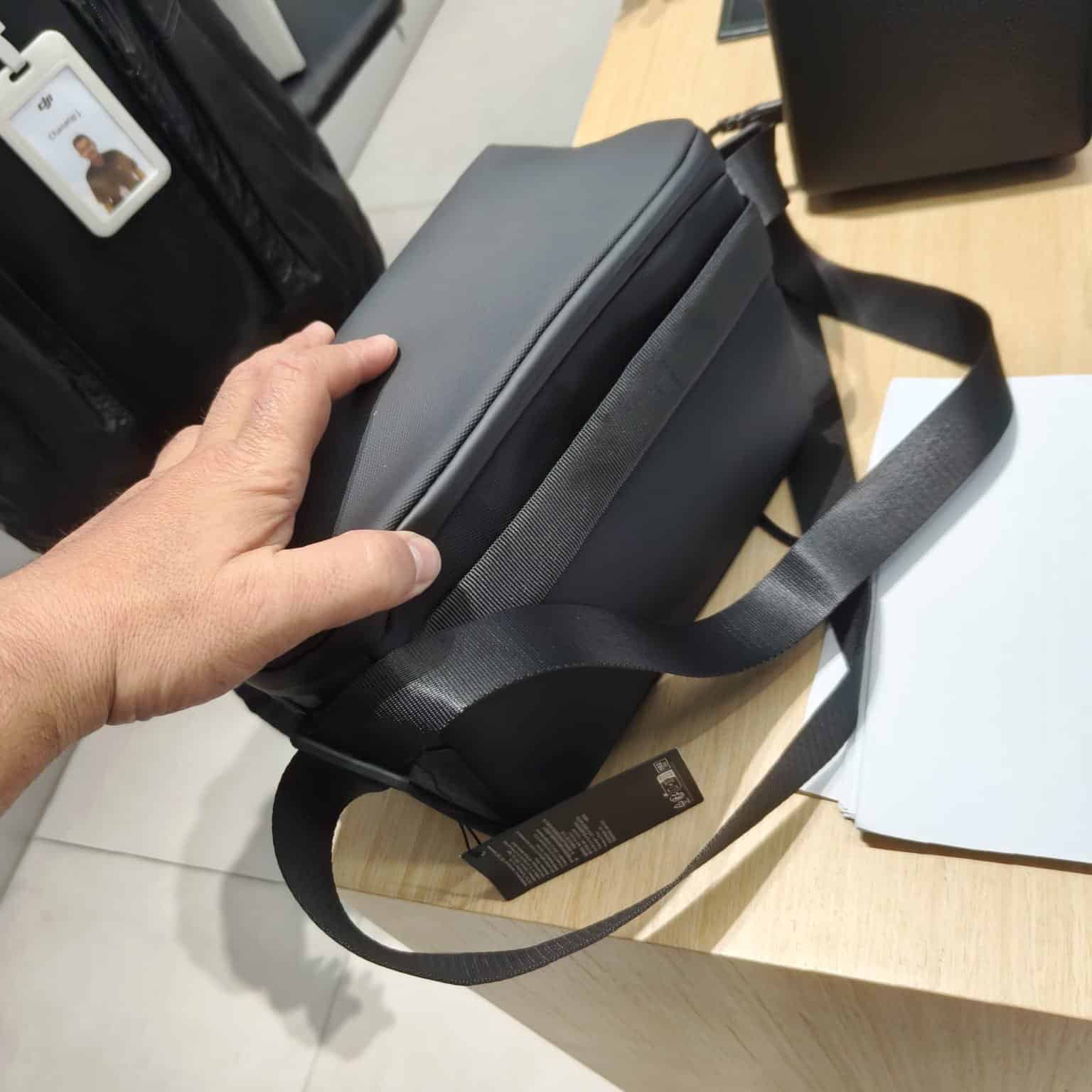 Hand lifting the closed grey shoulder bag by its strap on a desk.