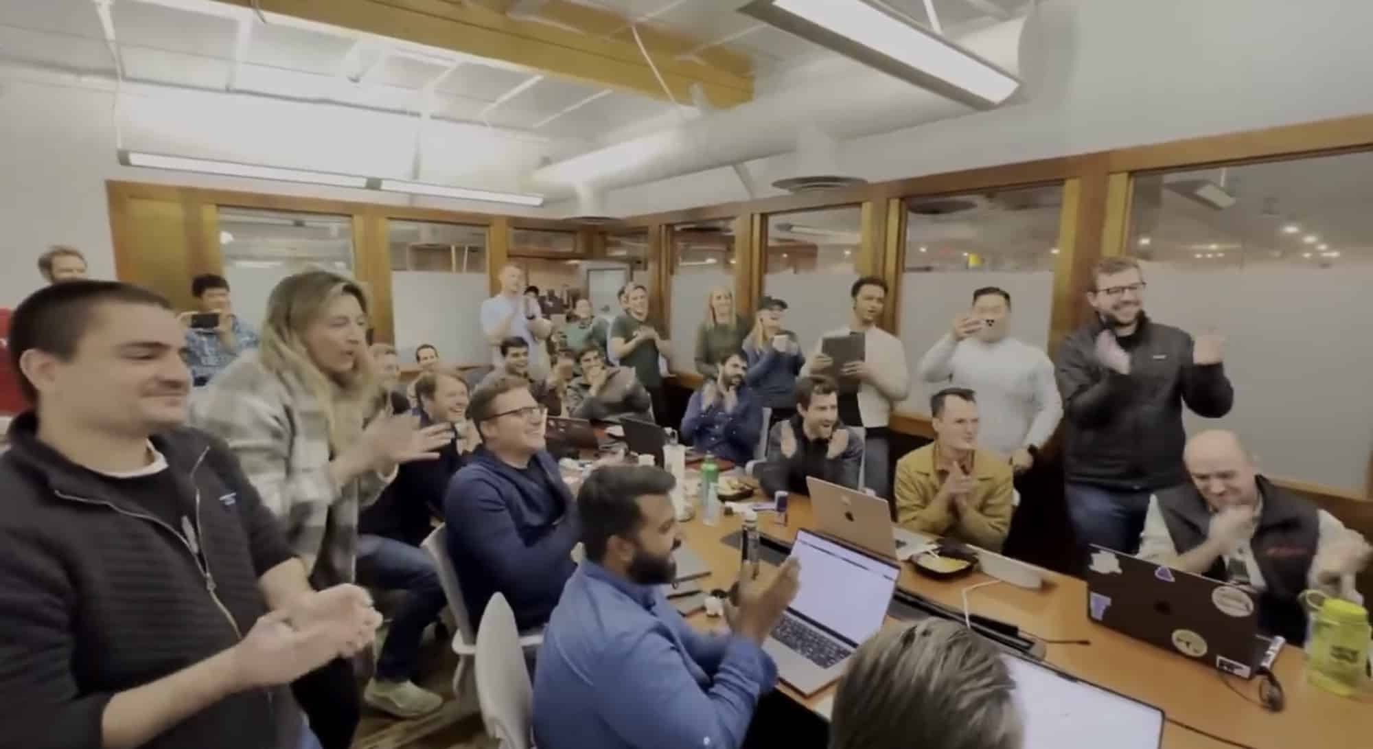 Office team applauds around a conference table filled with laptops.