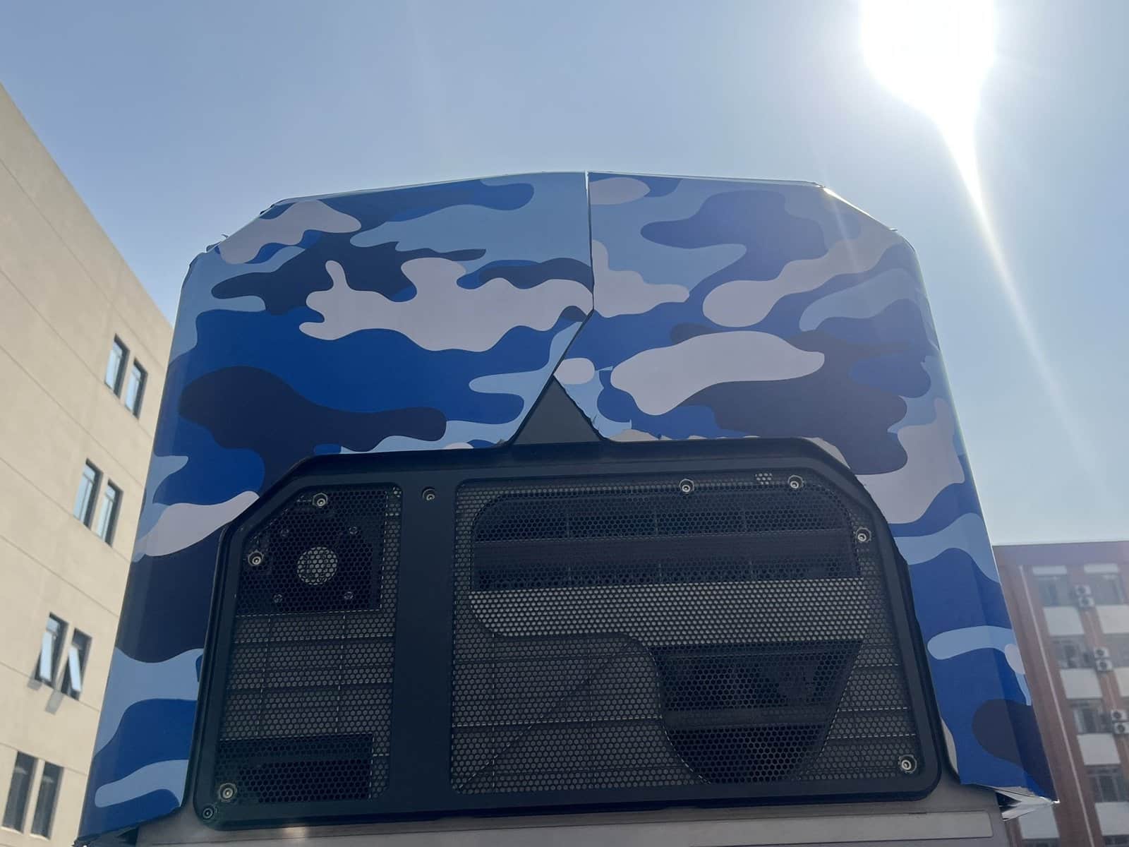 A blue camouflage-wrapped drone dock closed on a vehicle roof beside an office block.