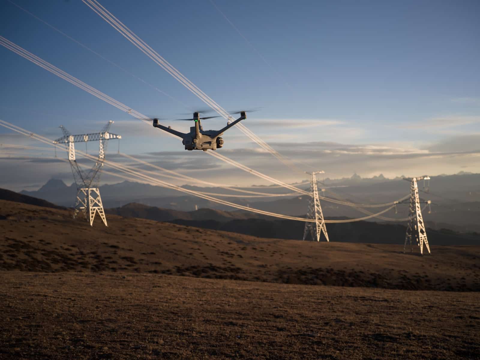 Single drone flying beneath sagging conductors between three transmission towers.