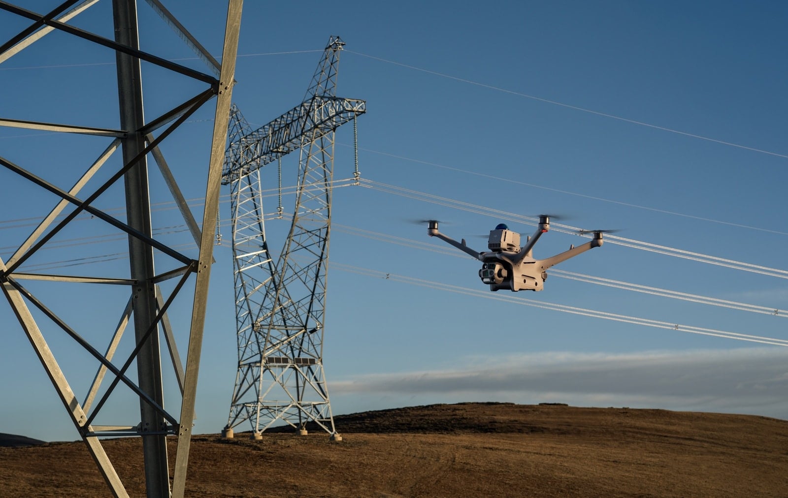 A tan DJI enterprise quadcopter flies past high-voltage transmission towers on a bare ridge.