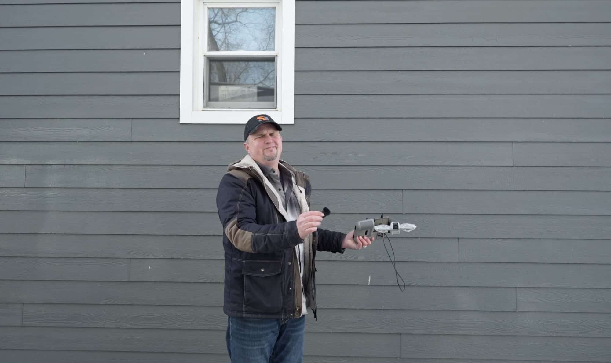 A reviewer holds a folded drone and a wireless microphone beside grey clapboard siding.