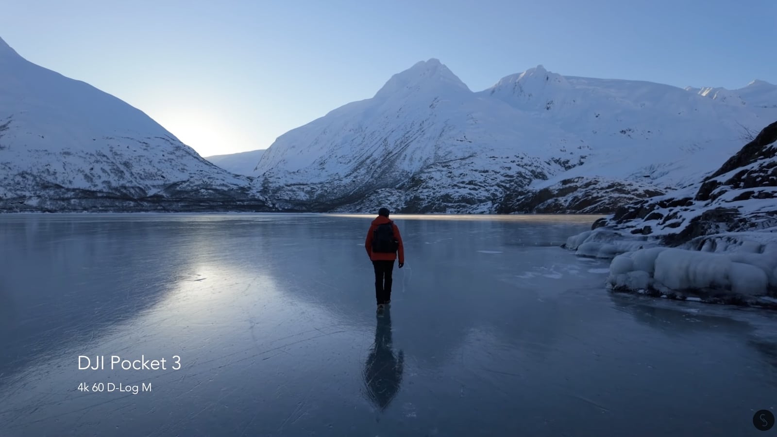 Lone figure in red walking across mirror-flat ice below snowy peaks.