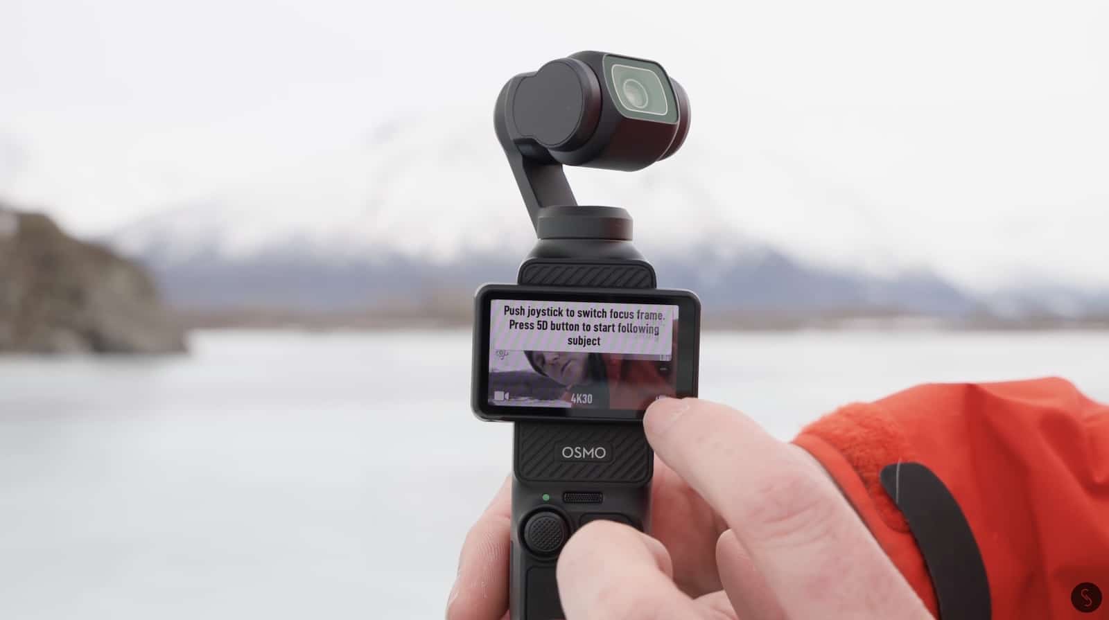 Hand holding the Osmo Pocket 3 on a frozen lake with an onscreen prompt.