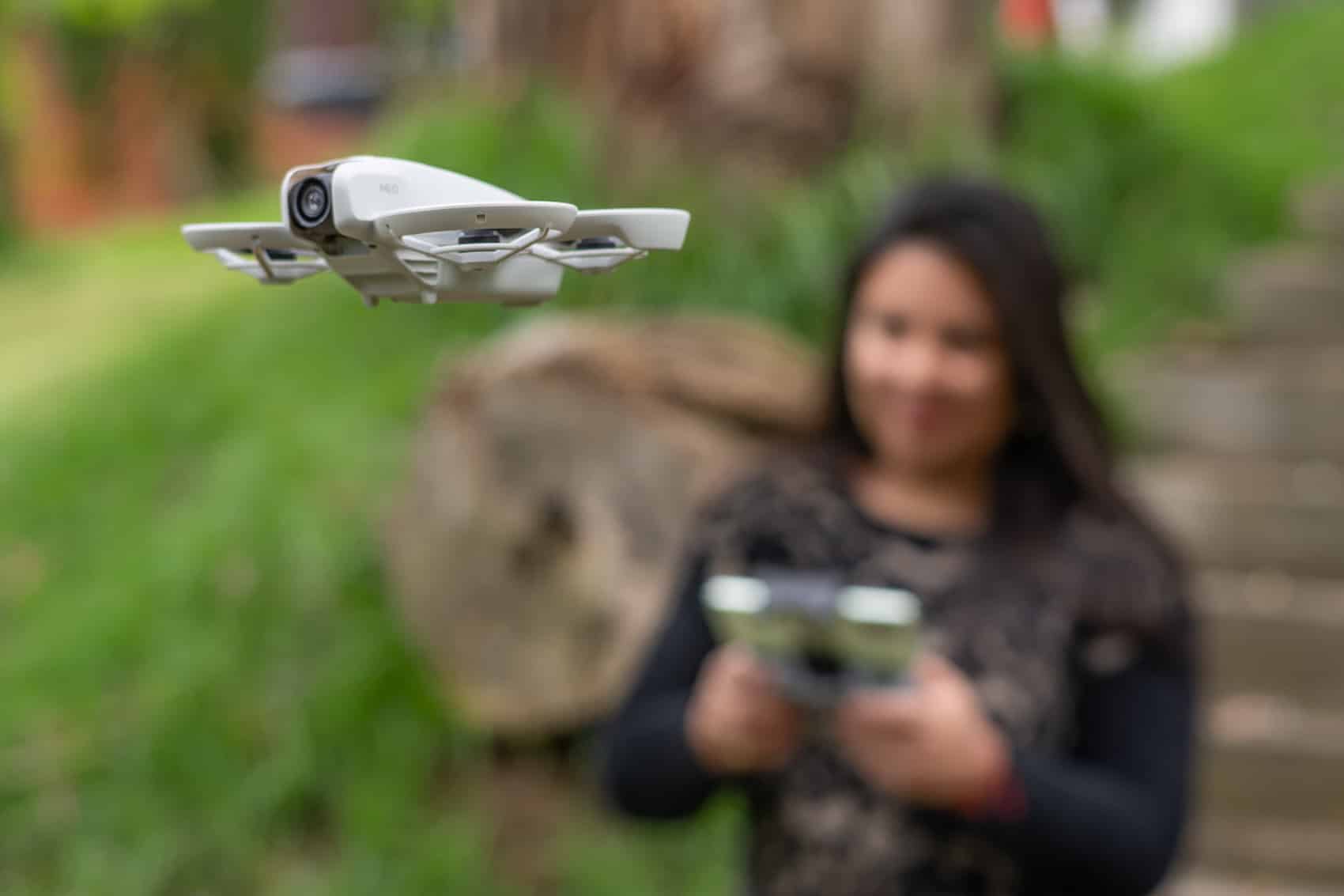 DJI Neo hovering in front of a woman holding a smartphone outdoors.