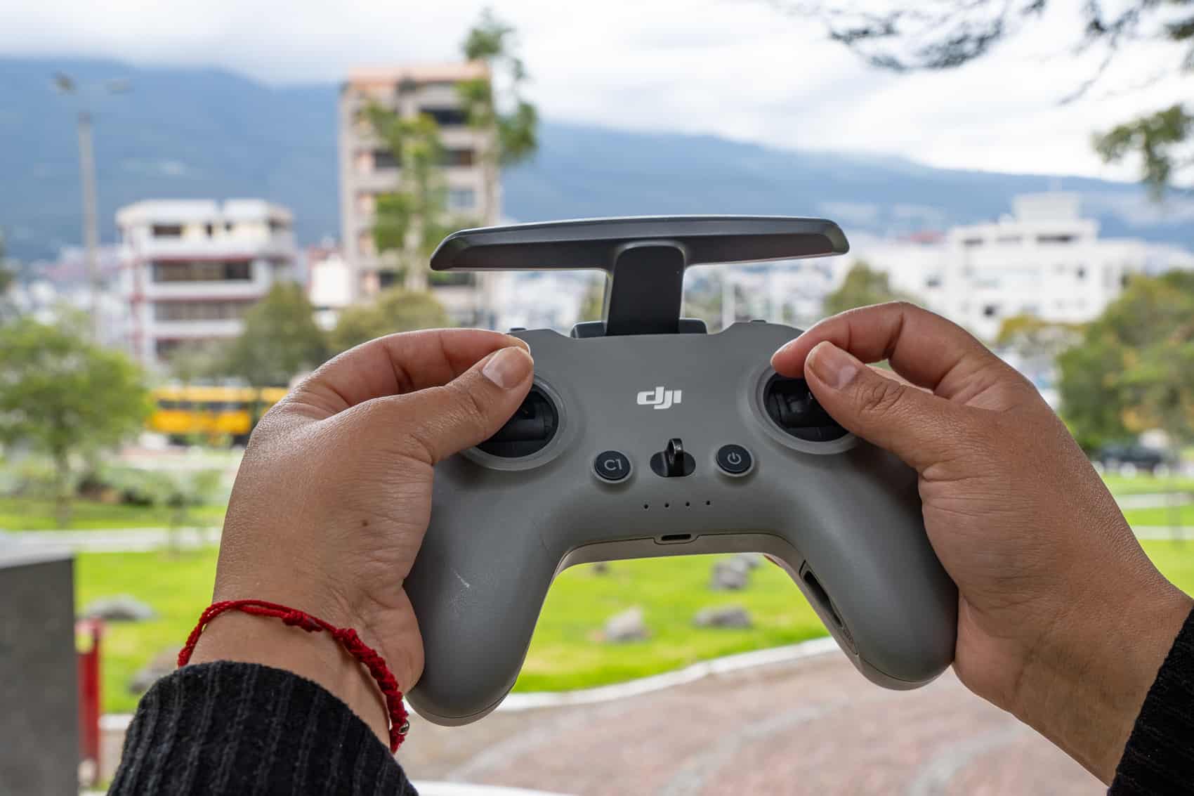 Hands grip a DJI remote controller outdoors with apartment blocks and green hills behind.