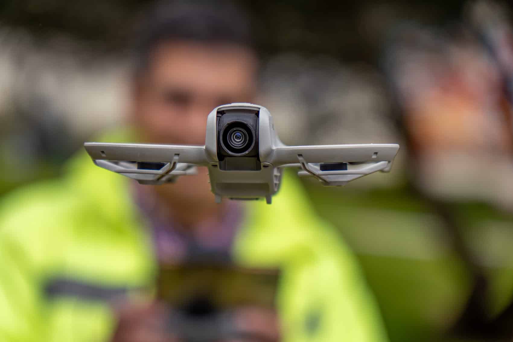 Small folding drone hovers in the foreground while a blurred pilot flies it.