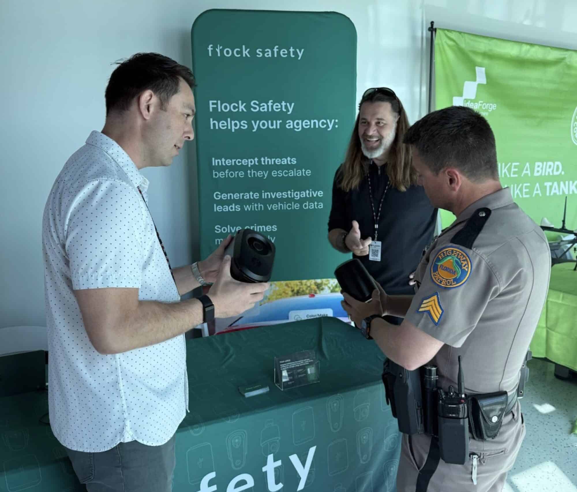 Two men and a woman talk beside a Flock Safety banner, one in uniform.