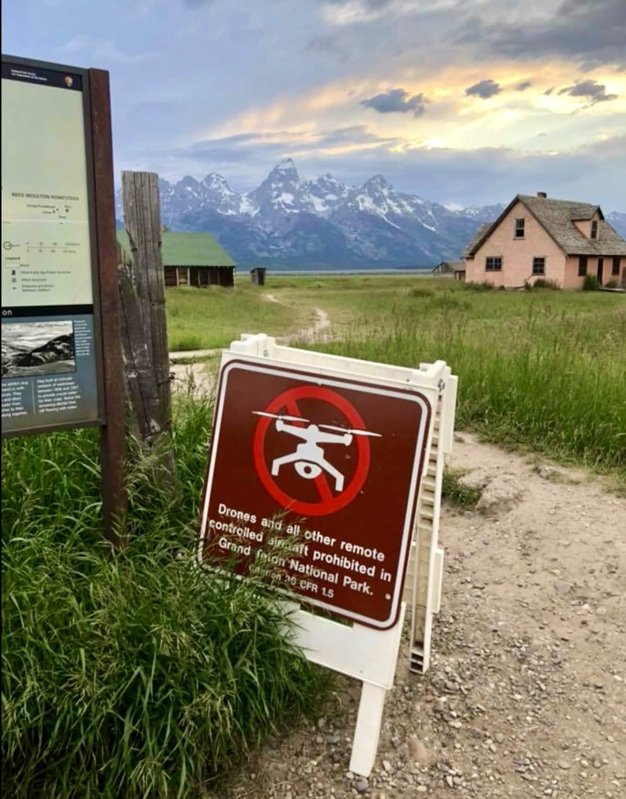 Trailhead sign banning drone launches stands before mountains and an old homestead.