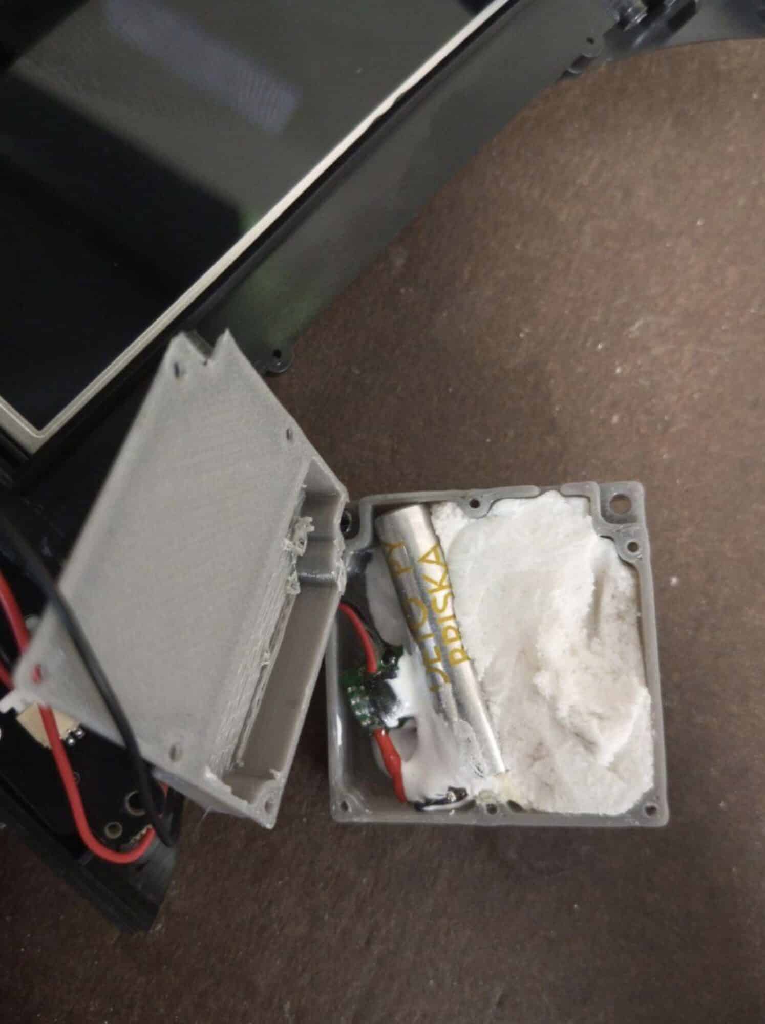 Opened gray housing on a floor packed with white putty and wired battery leads.