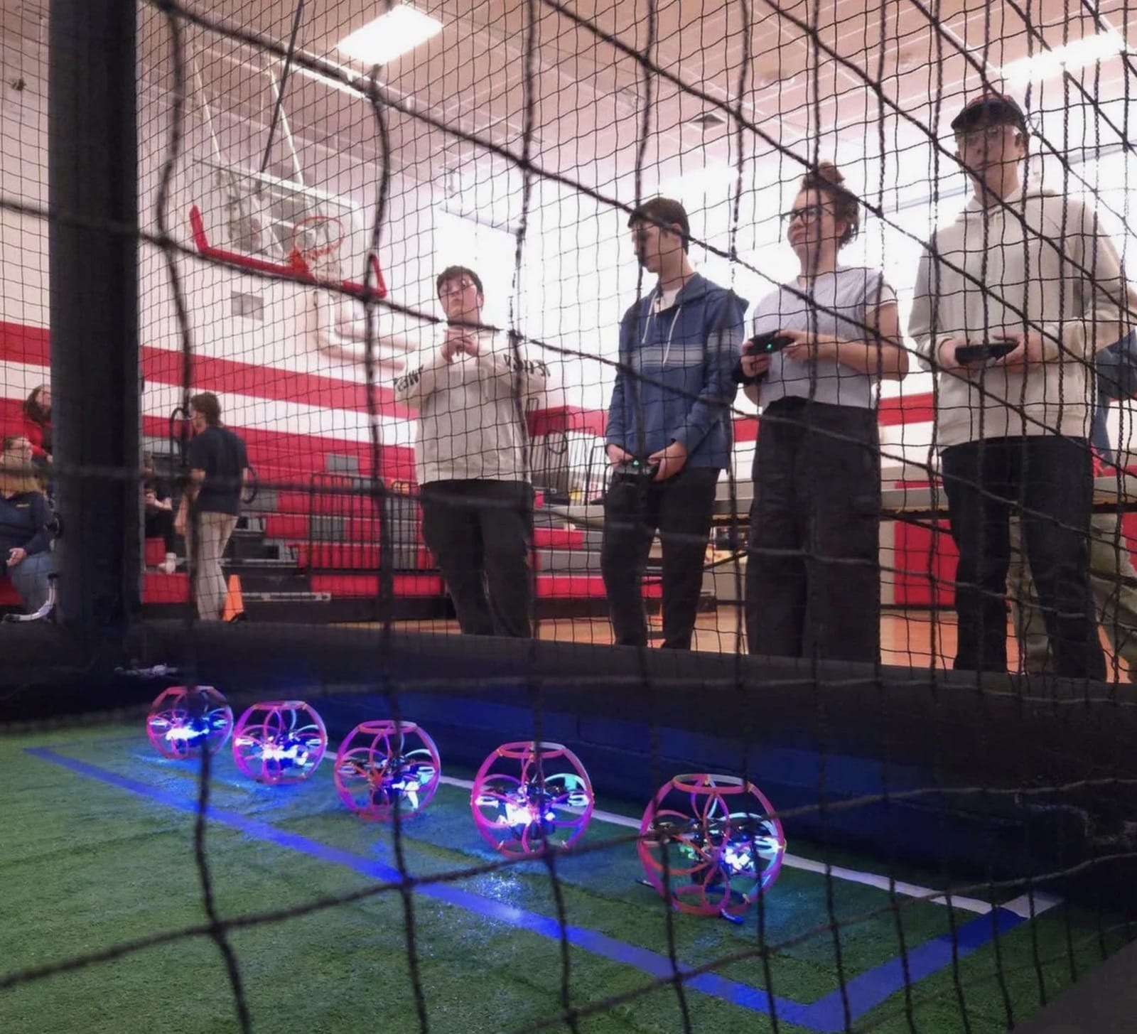 Students pilot glowing drone soccer balls inside a netted indoor arena.