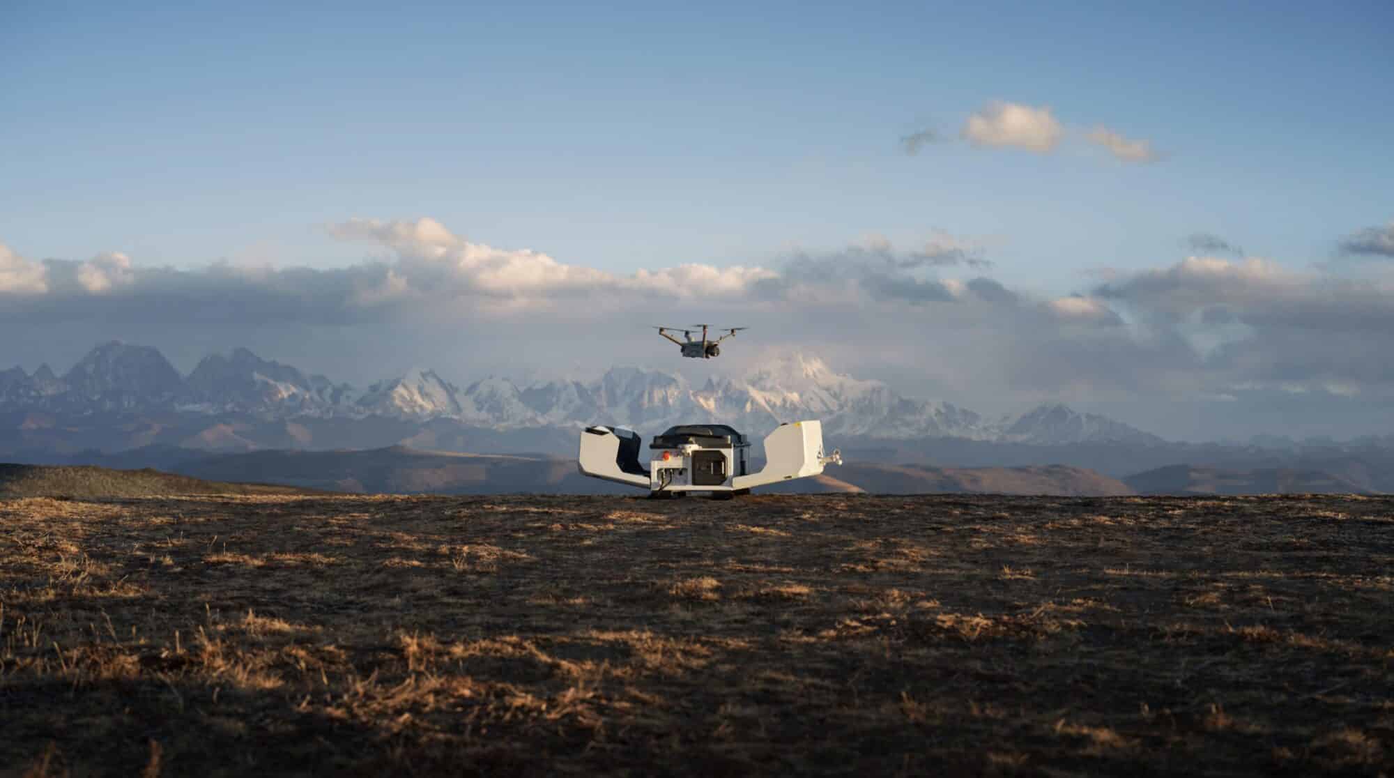 Drone hovering above an open dock on a plateau below snowy peaks.