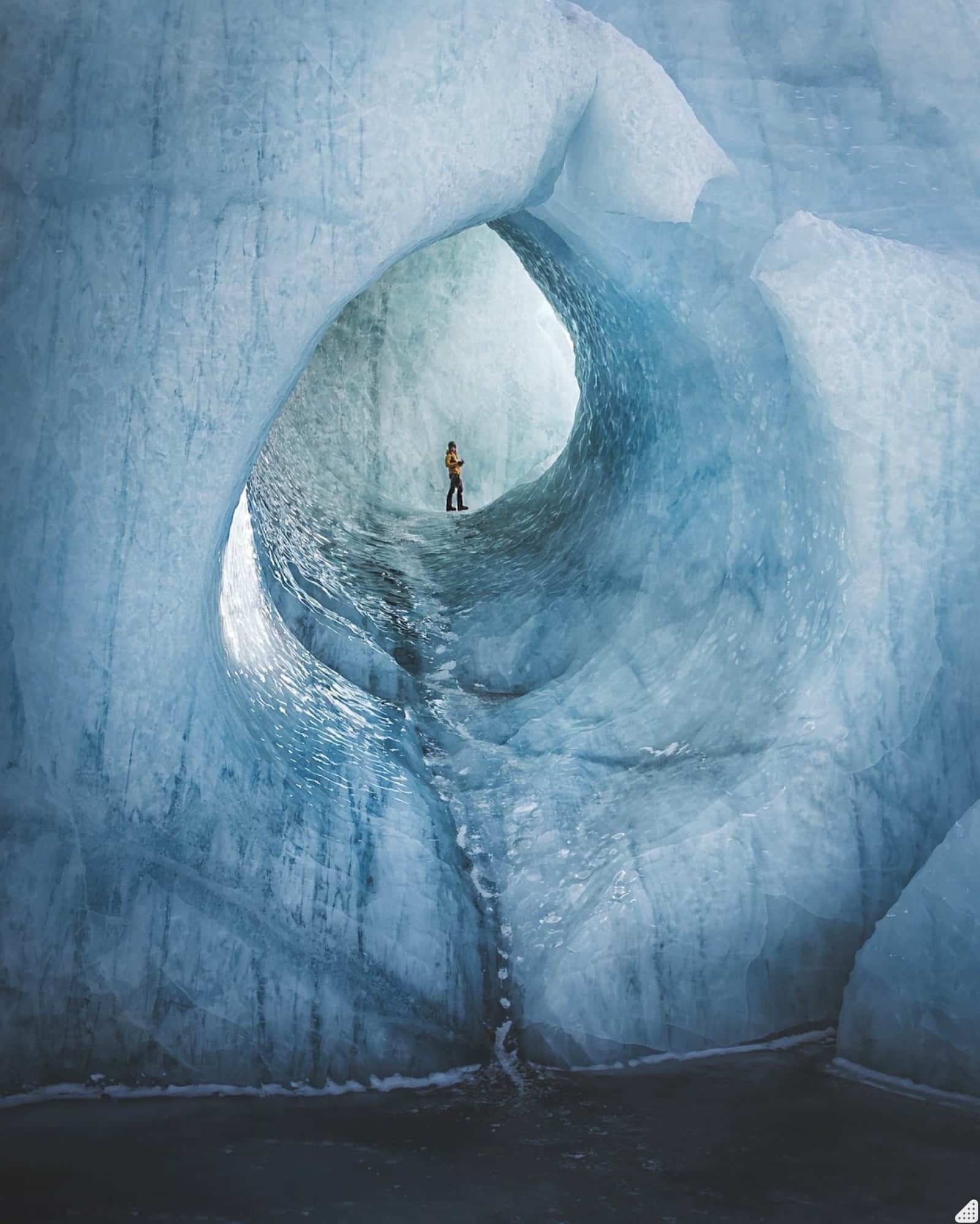 A tiny figure stands inside a cavernous blue glacier ice tunnel.