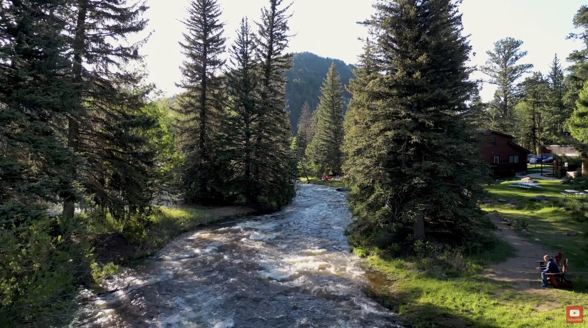 Aerial view of a rushing mountain stream winding through pines past small cabins.