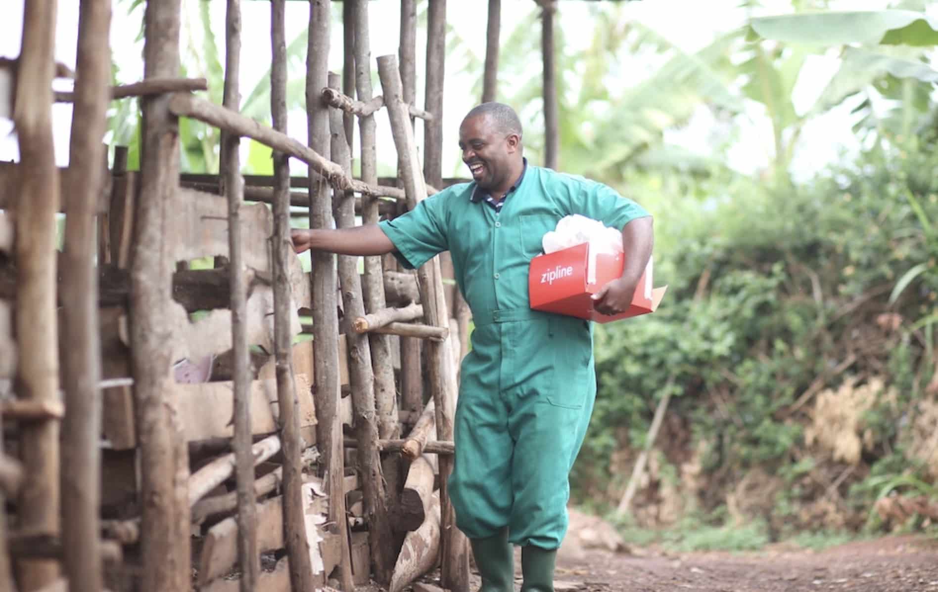 A worker in teal coveralls carries a Zipline delivery box past a wooden livestock pen.