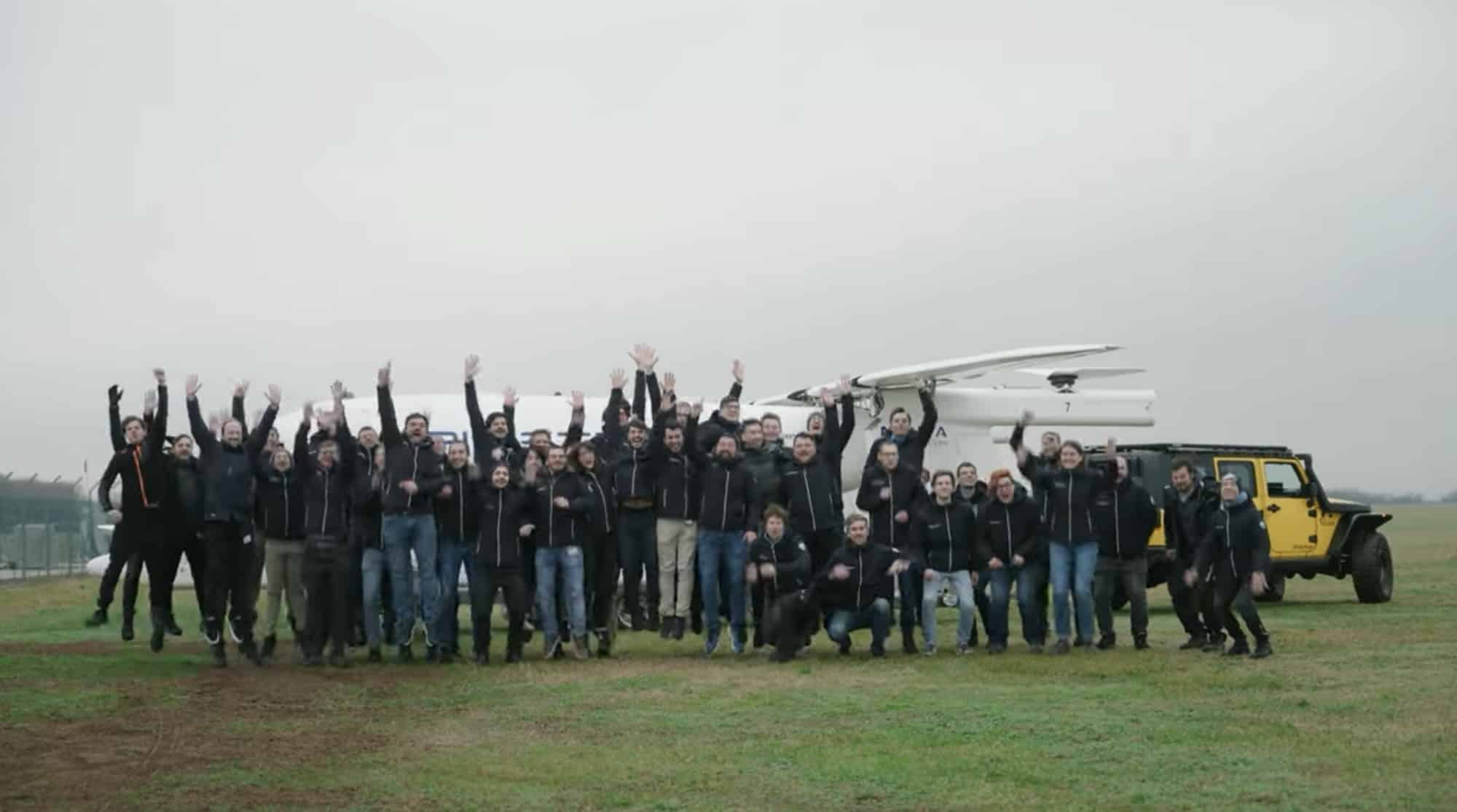 The development team leaps in celebration on grass beside the aircraft.