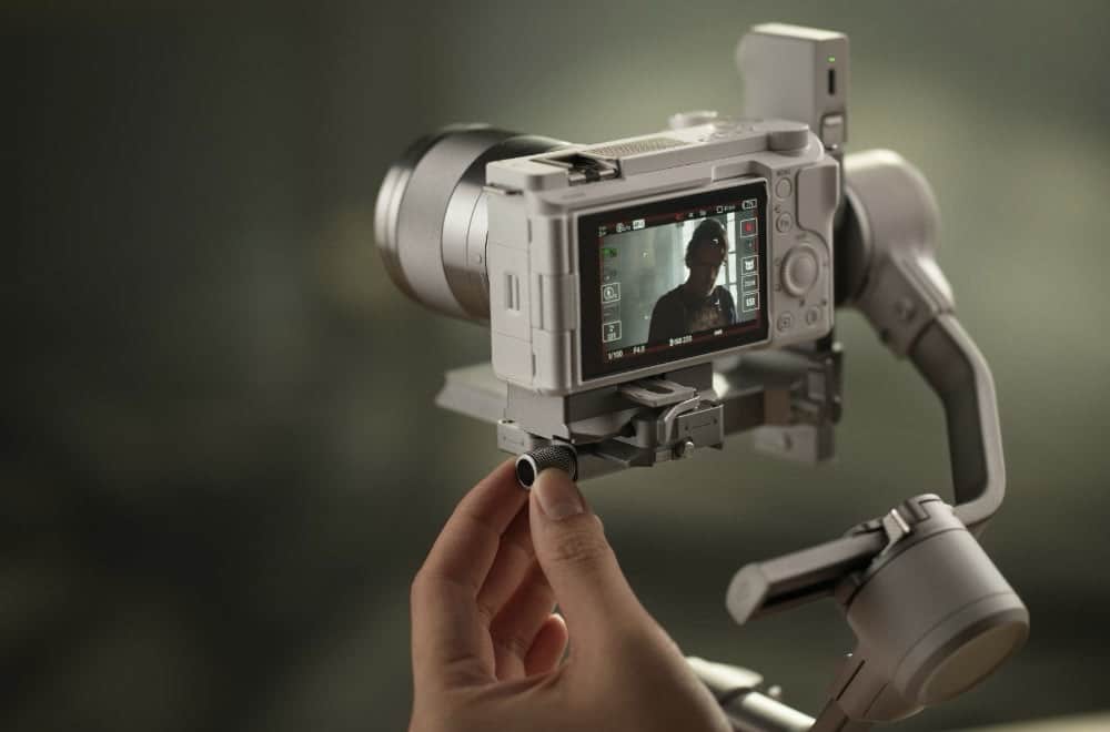 A hand adjusts a compact mirrorless camera balanced on the white gimbal.