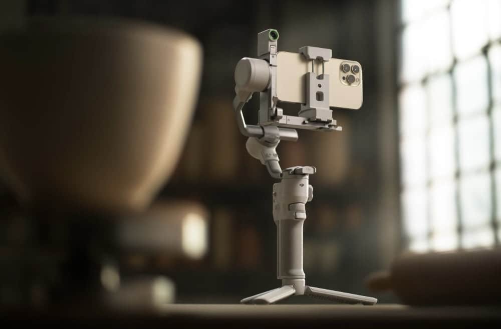 The gimbal holds a smartphone on its mini tripod beside a ceramic bowl.