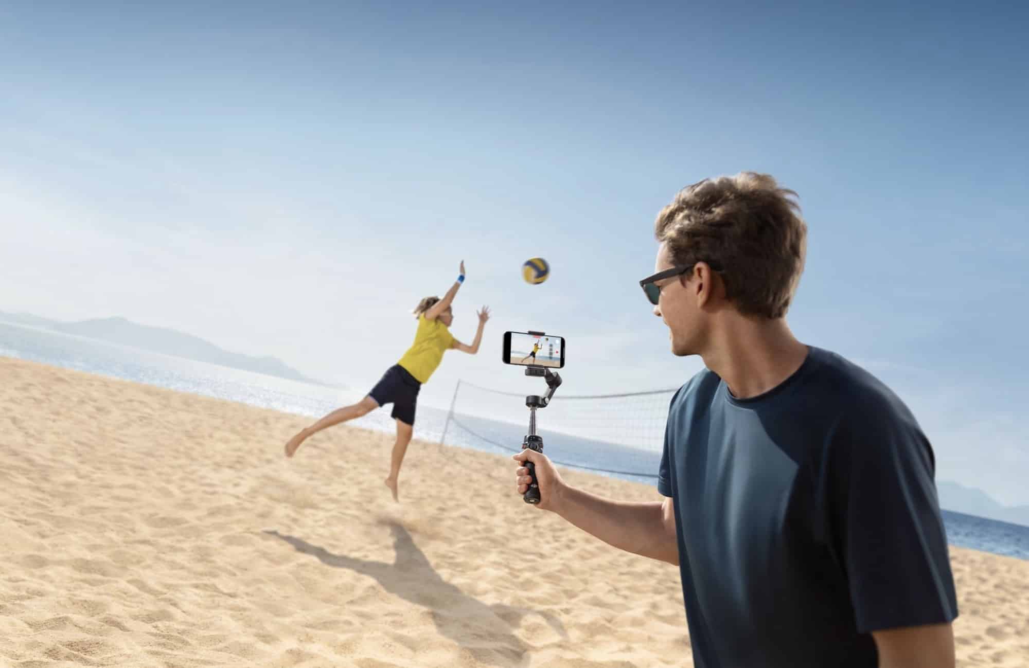 A man on a beach films a friend leaping for a ball with a phone gimbal.