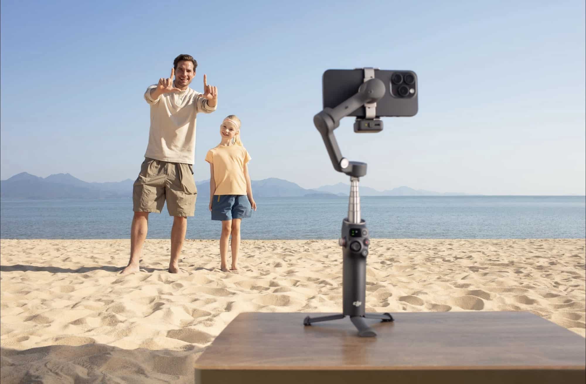 A man and child wave at the tripod-mounted gimbal filming them on the sand.