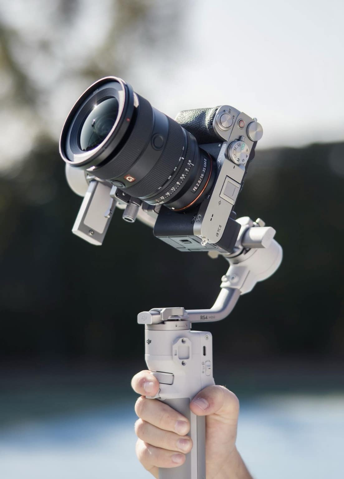 A hand holds the gimbal with a Sony camera and wide zoom beside water.