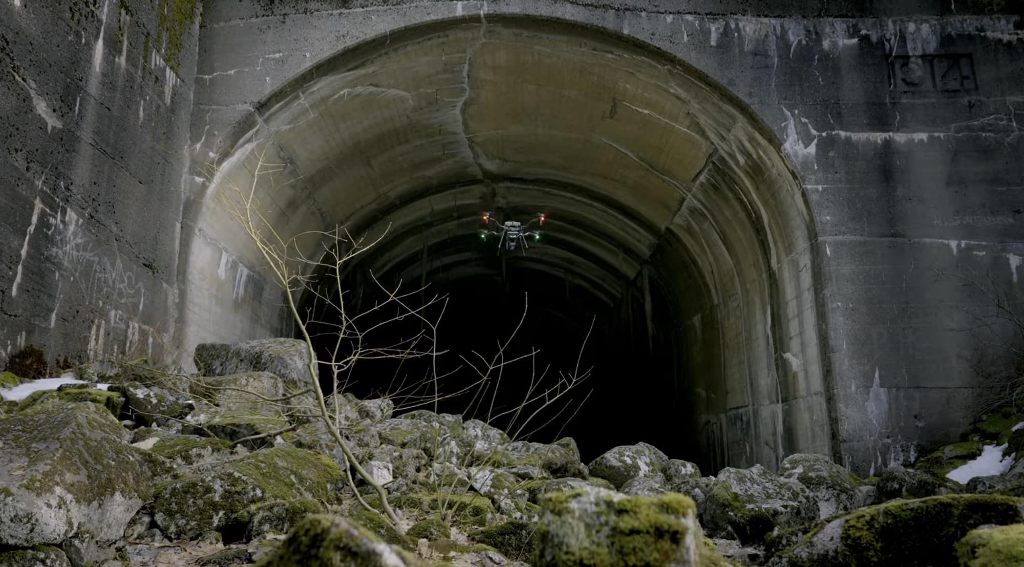 A quadcopter hovers at the mouth of a dark concrete tunnel above mossy rocks.