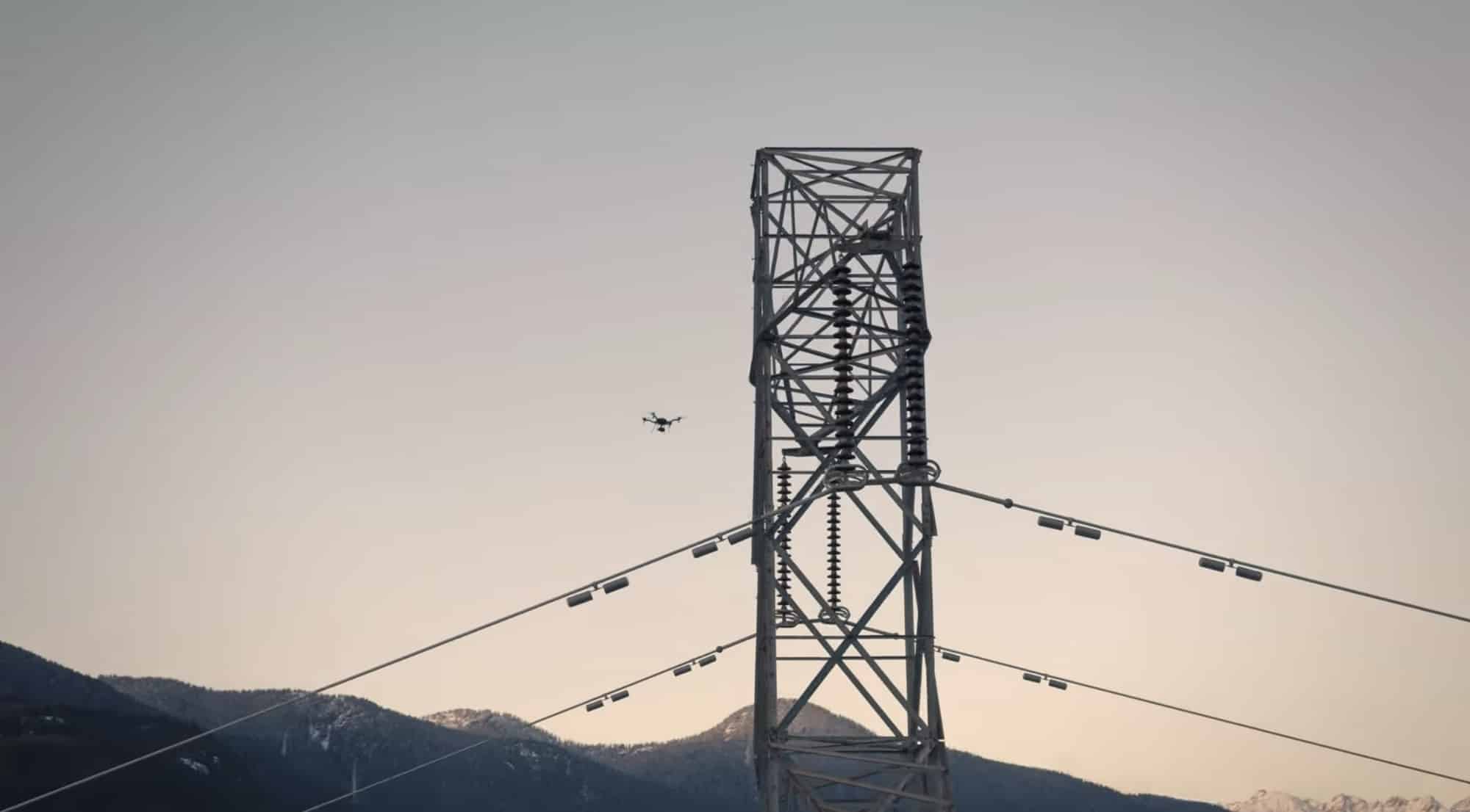 A drone inspects a high-voltage transmission tower at dusk over mountain ridges.