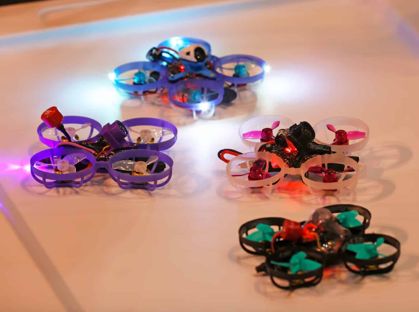 Several tiny ducted racing quadcopters glow with coloured LEDs on a table.