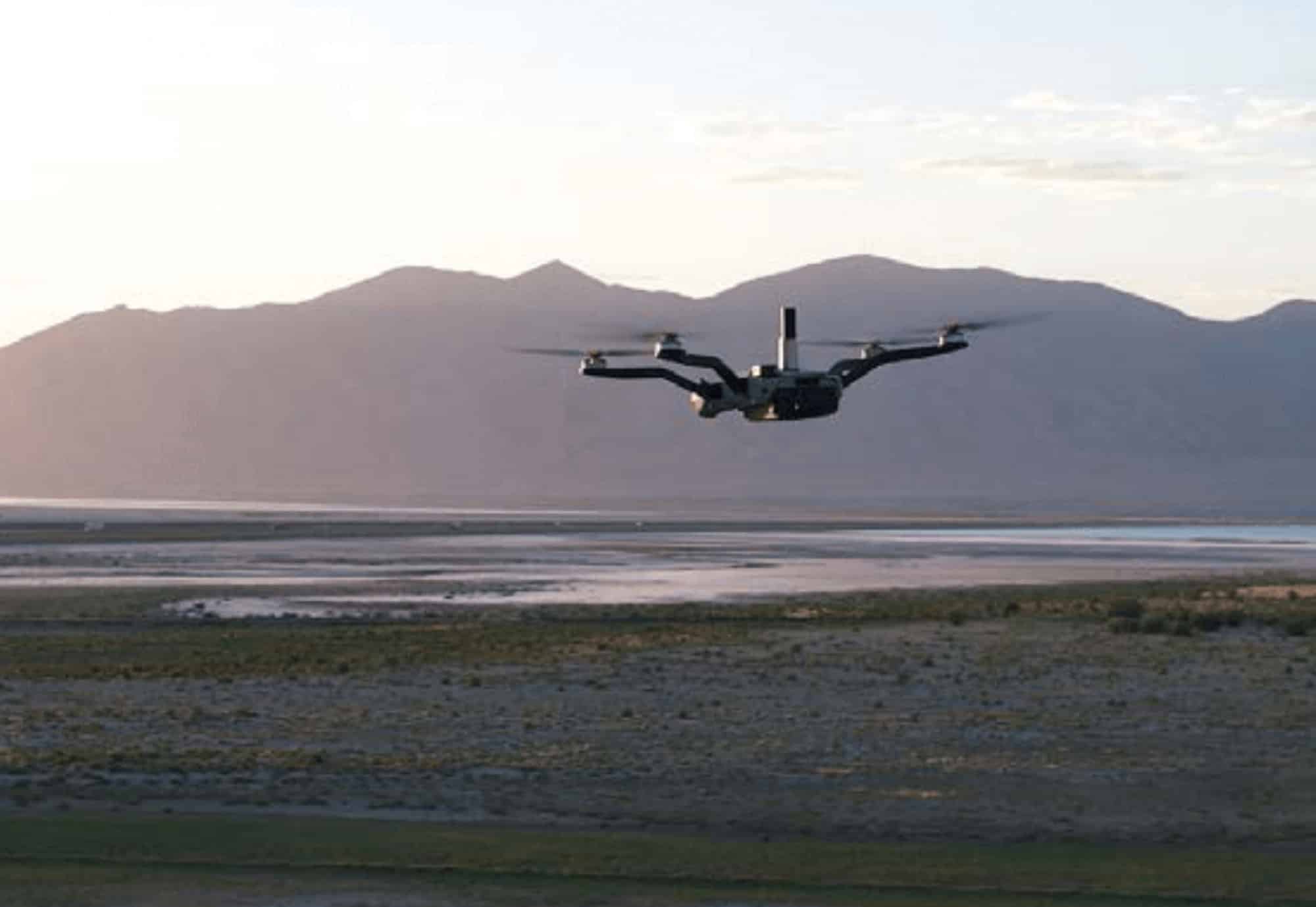 Black quadcopter hovering above a shallow salt flat below bare mountains.