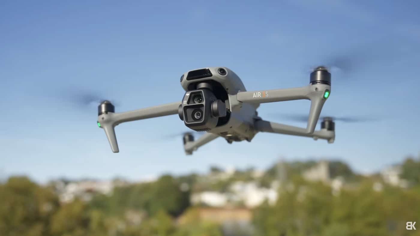 A DJI Air 3S hovers against a clear blue sky above distant treetops.
