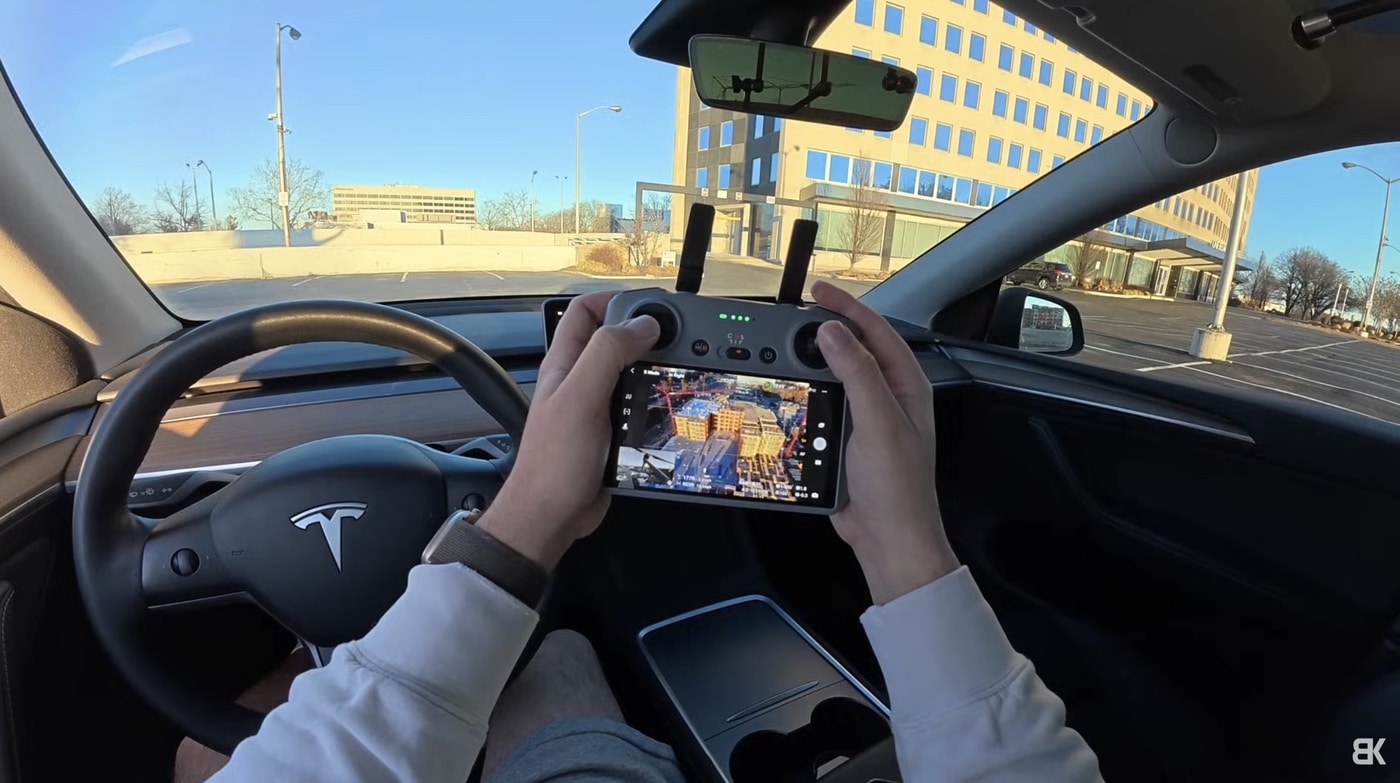 Hands work a drone controller over the steering wheel of a parked Tesla.