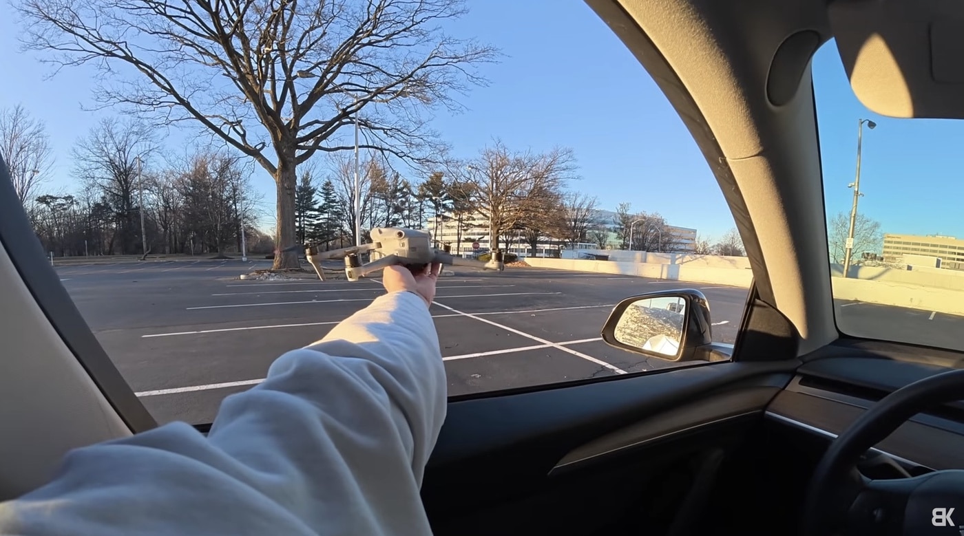 A pilot reaches out of a car window holding a folded drone over empty parking spaces.